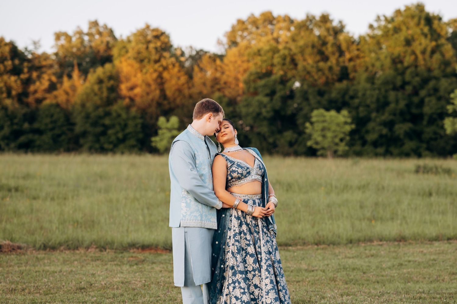 Couple in formal attire embracing in a green field with golden autumn trees at sunset.