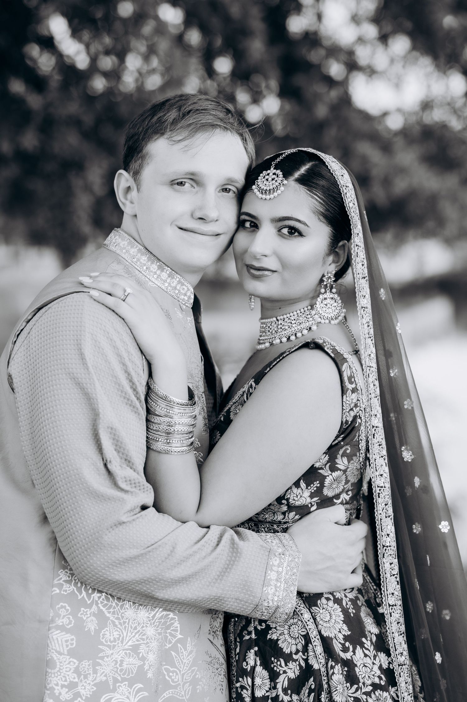 Black and white portrait of a couple in traditional South Asian wedding attire, embracing and smiling warmly.