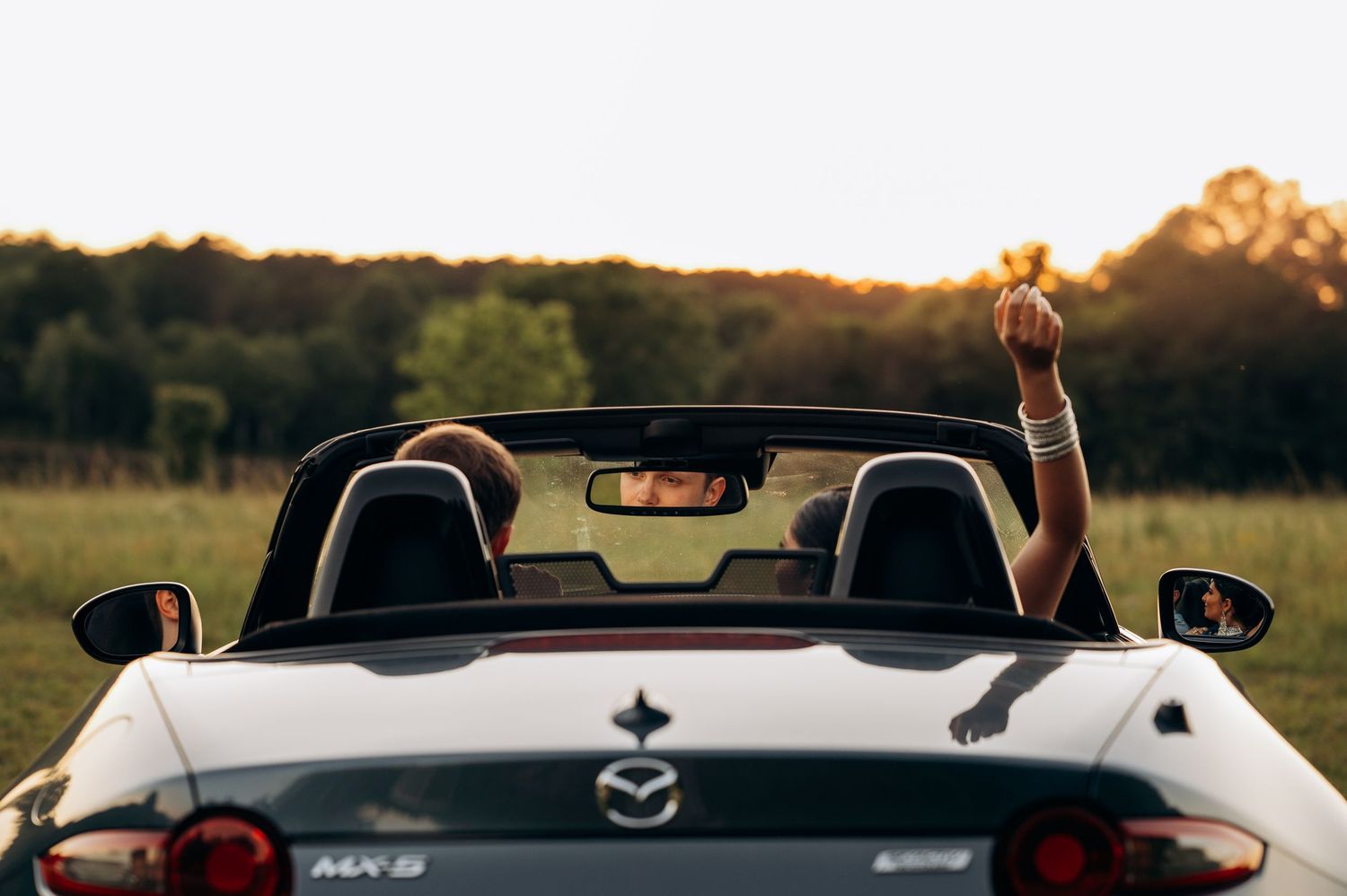 Person relaxing in a white Mazda MX-5 convertible at sunset in a green field, arm raised joyfully.