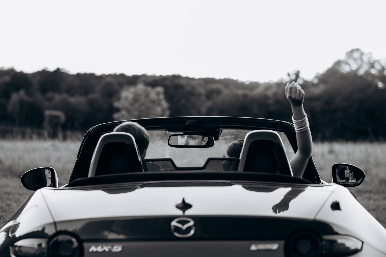 Black and white photo of a Mazda MX-5 convertible in a field, with a hand raised above the windshield.