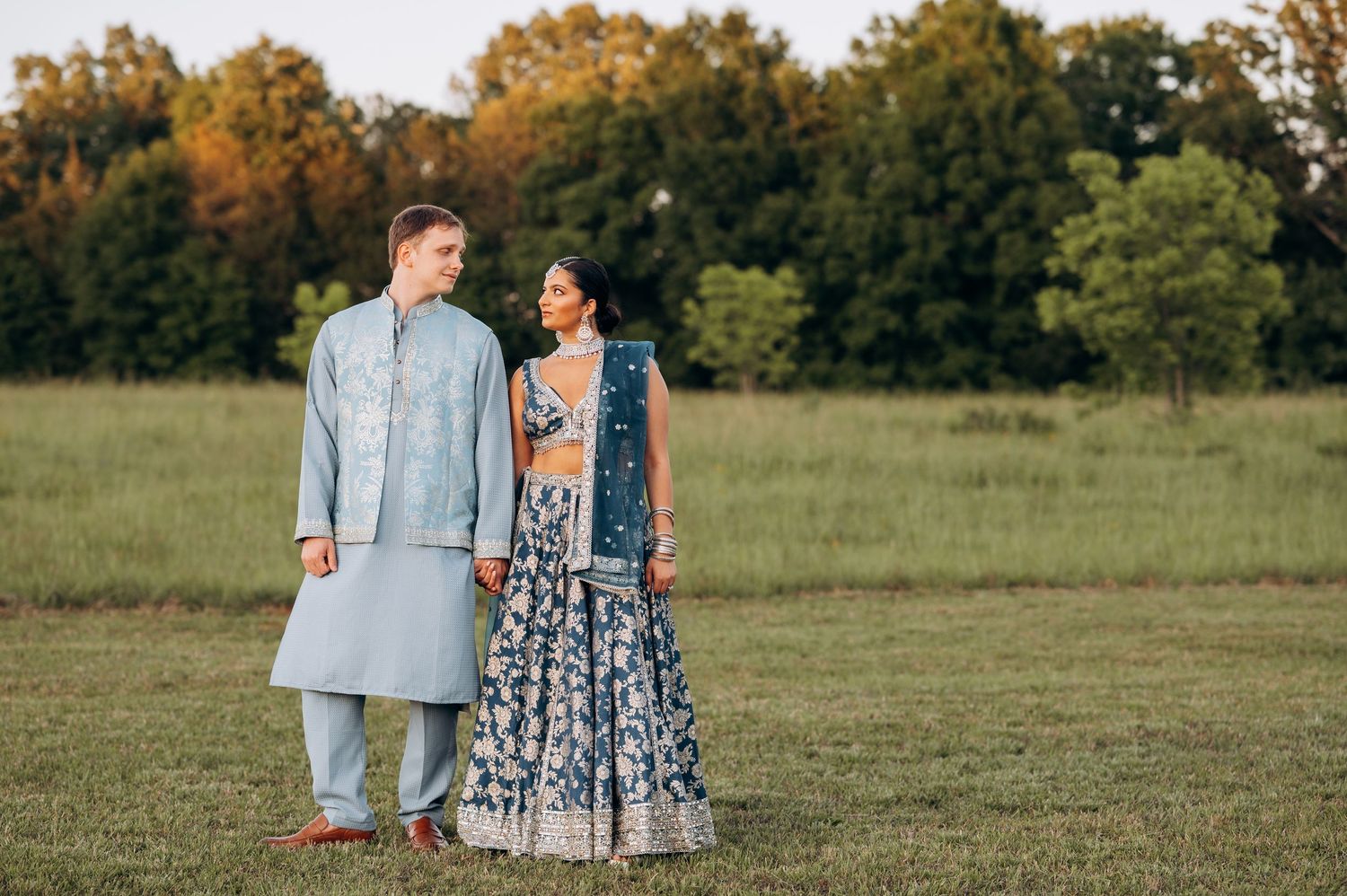 Couple in traditional Indian wedding attire holding hands in a green field, wearing blue lehenga and kurta.