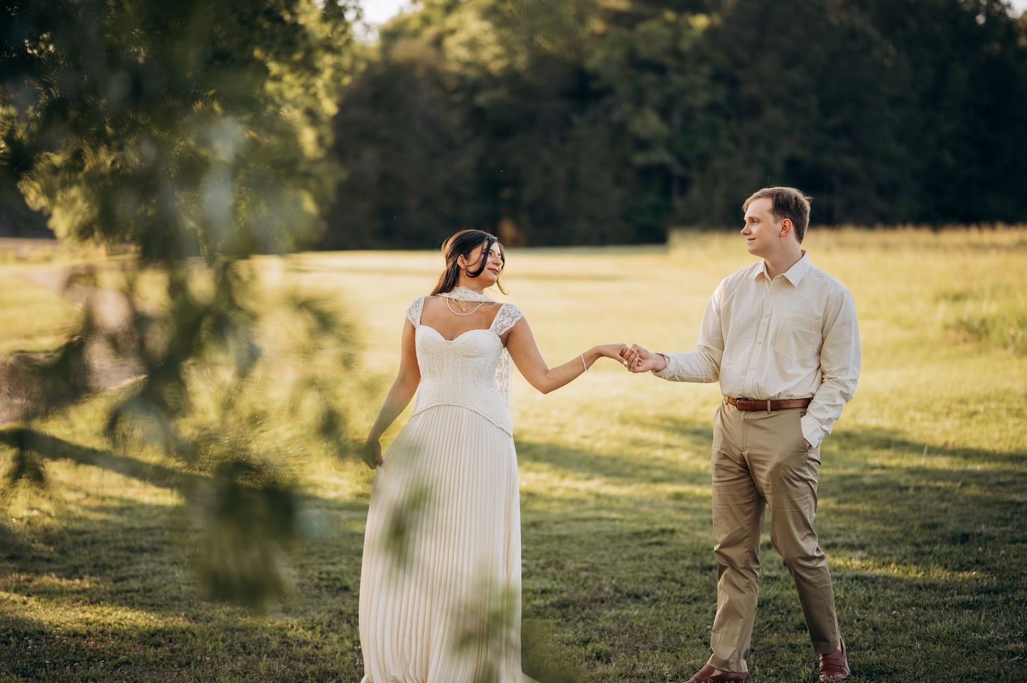 Bride and groom holding hands in a lush green field, she wears a flowing white gown with floral crown.