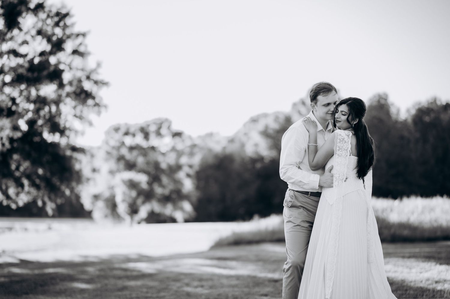 Black and white wedding photo of a couple embracing outdoors, groom kissing bride in white gown near lush trees.