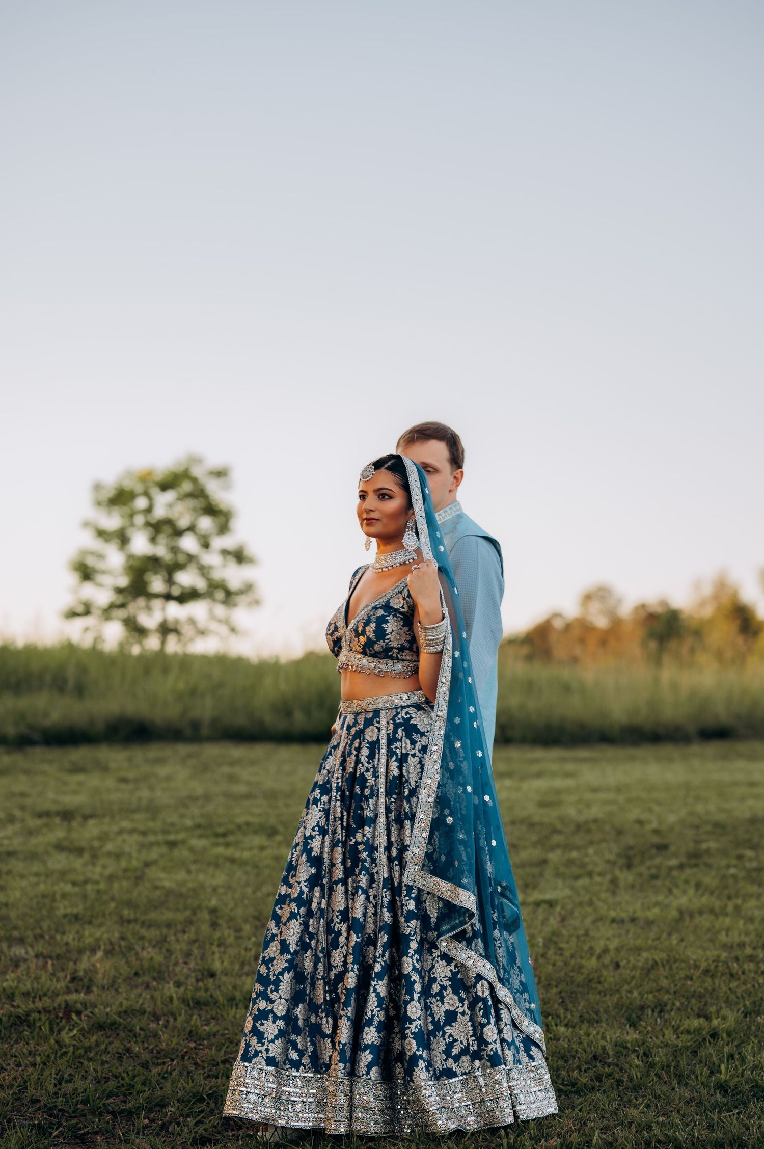 Couple in traditional Indian blue lehenga and kurta posing outdoors in a green field at dusk.