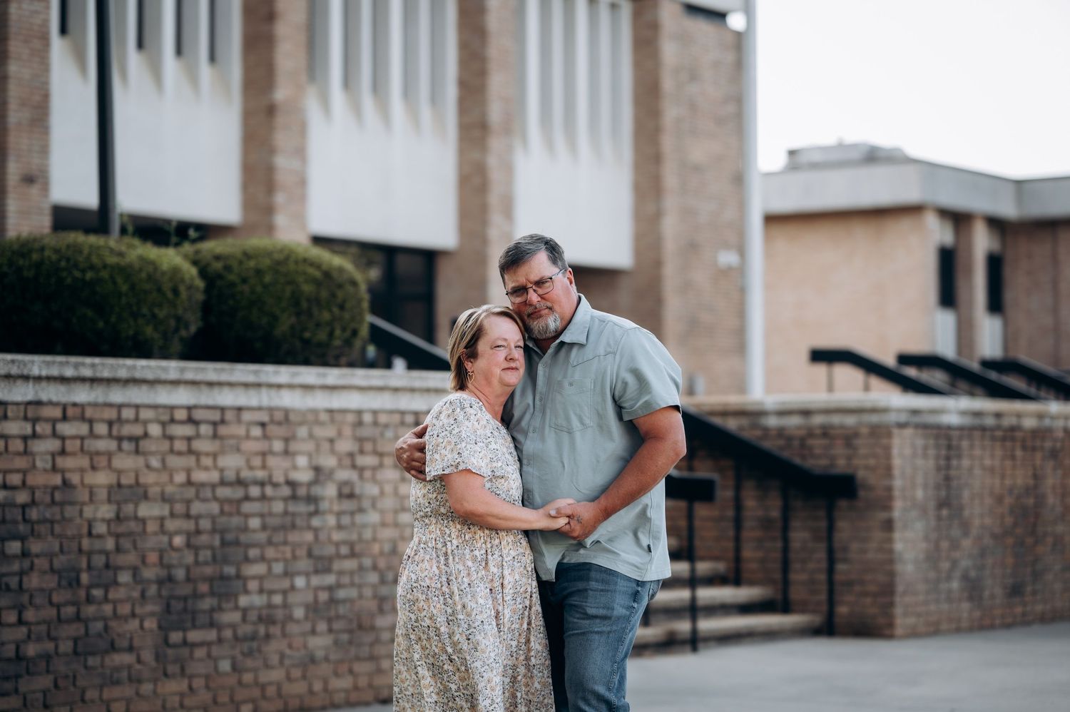 A couple embraces warmly outside a brick church building, sharing a tender moment together.