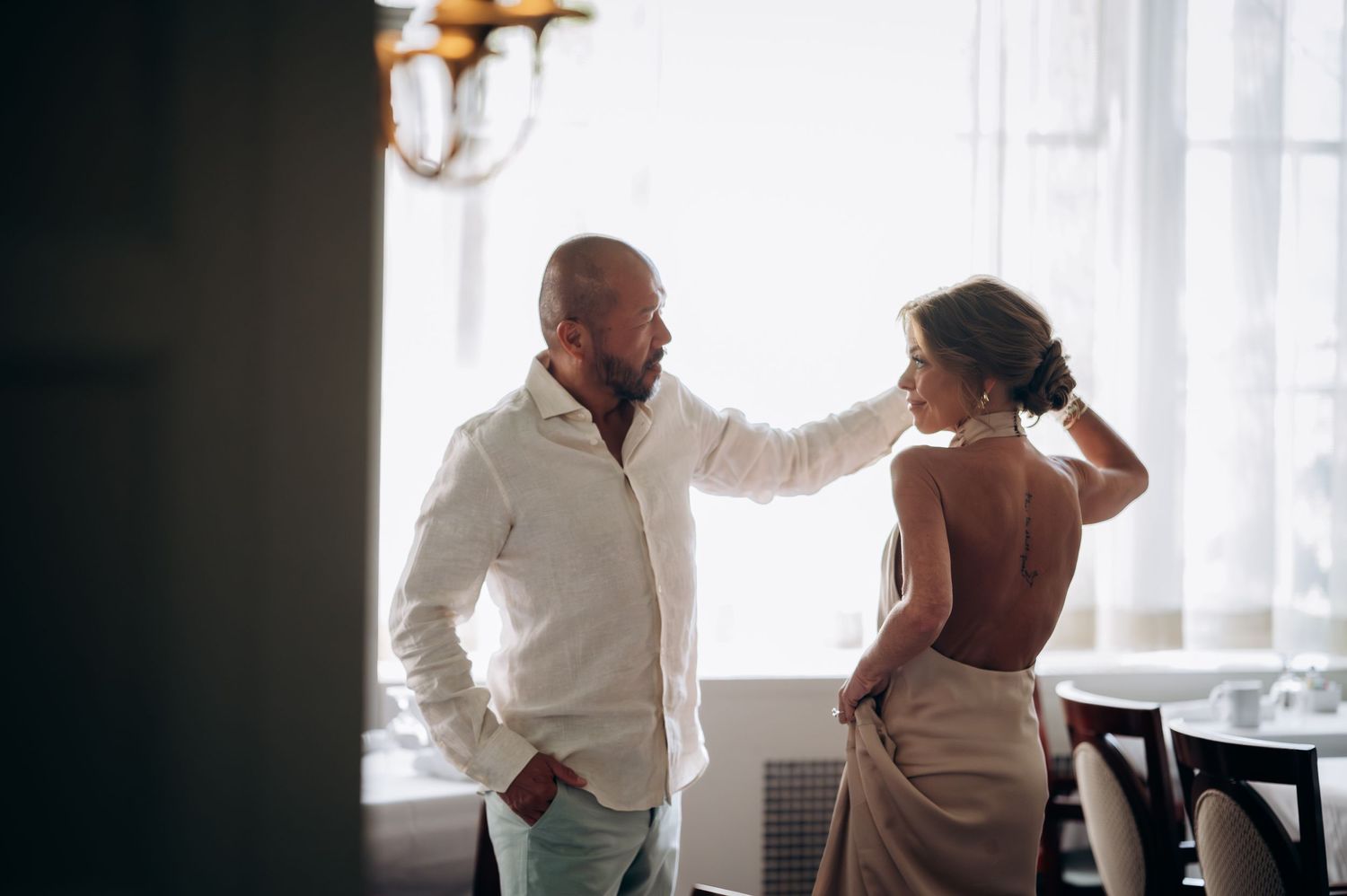 A bald man in a white shirt smiles at a woman in a backless dress near a bright window indoors.