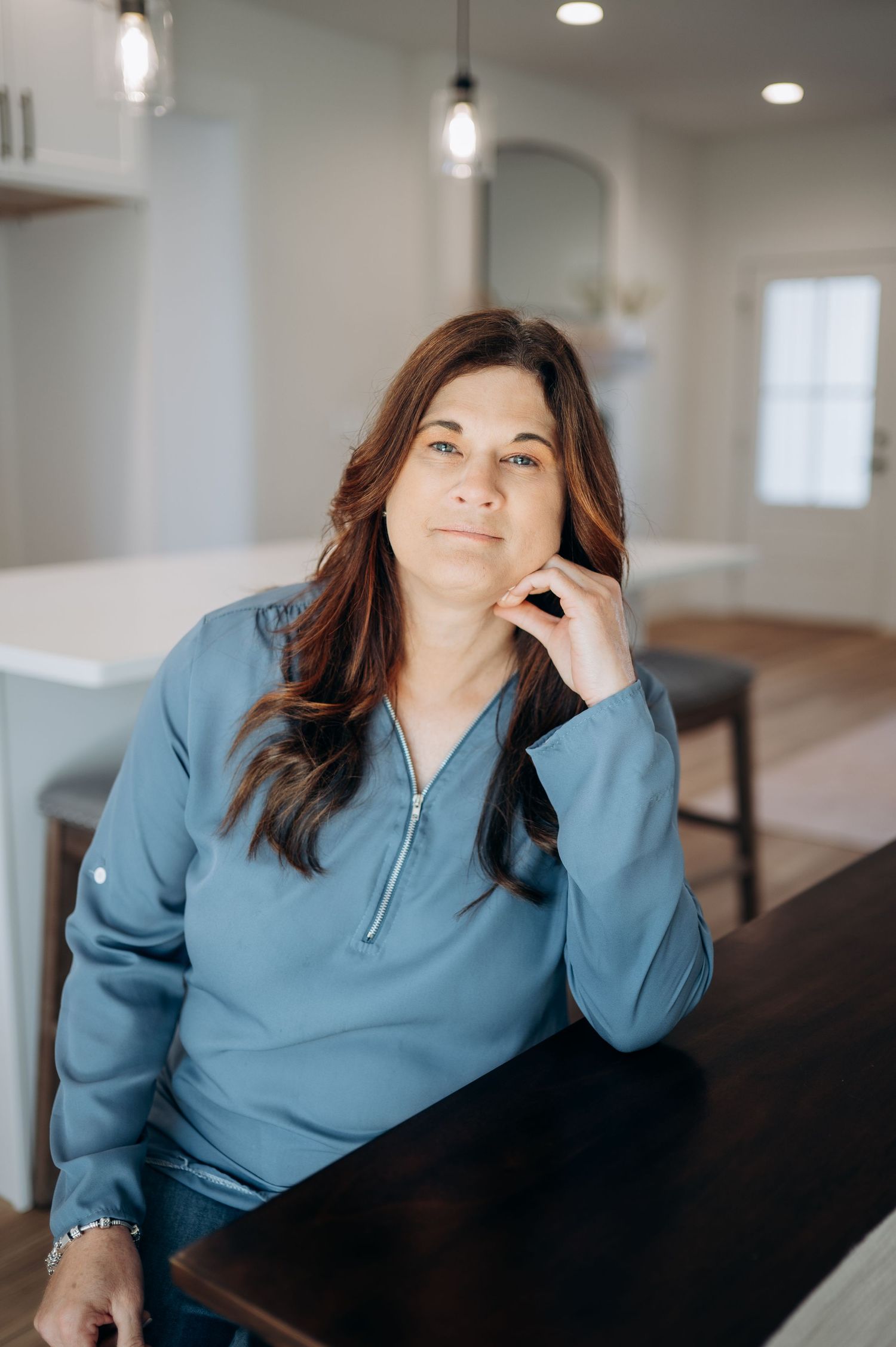 A woman with brown hair in a blue quarter-zip pullover sits thoughtfully at a dark wood table in a modern kitchen.