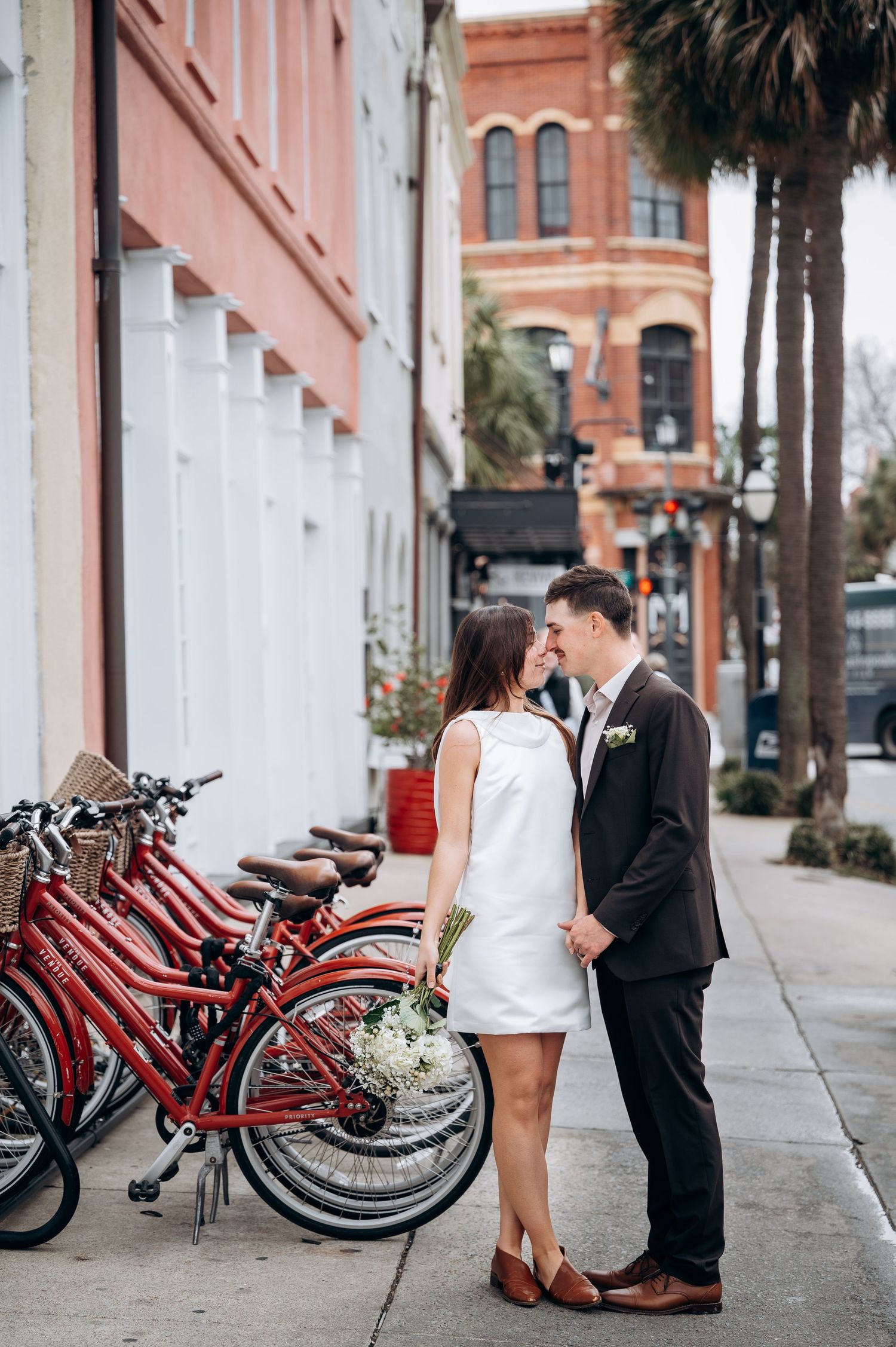 Couple in formal wear sharing romantic moment near red rental bikes on historic street.