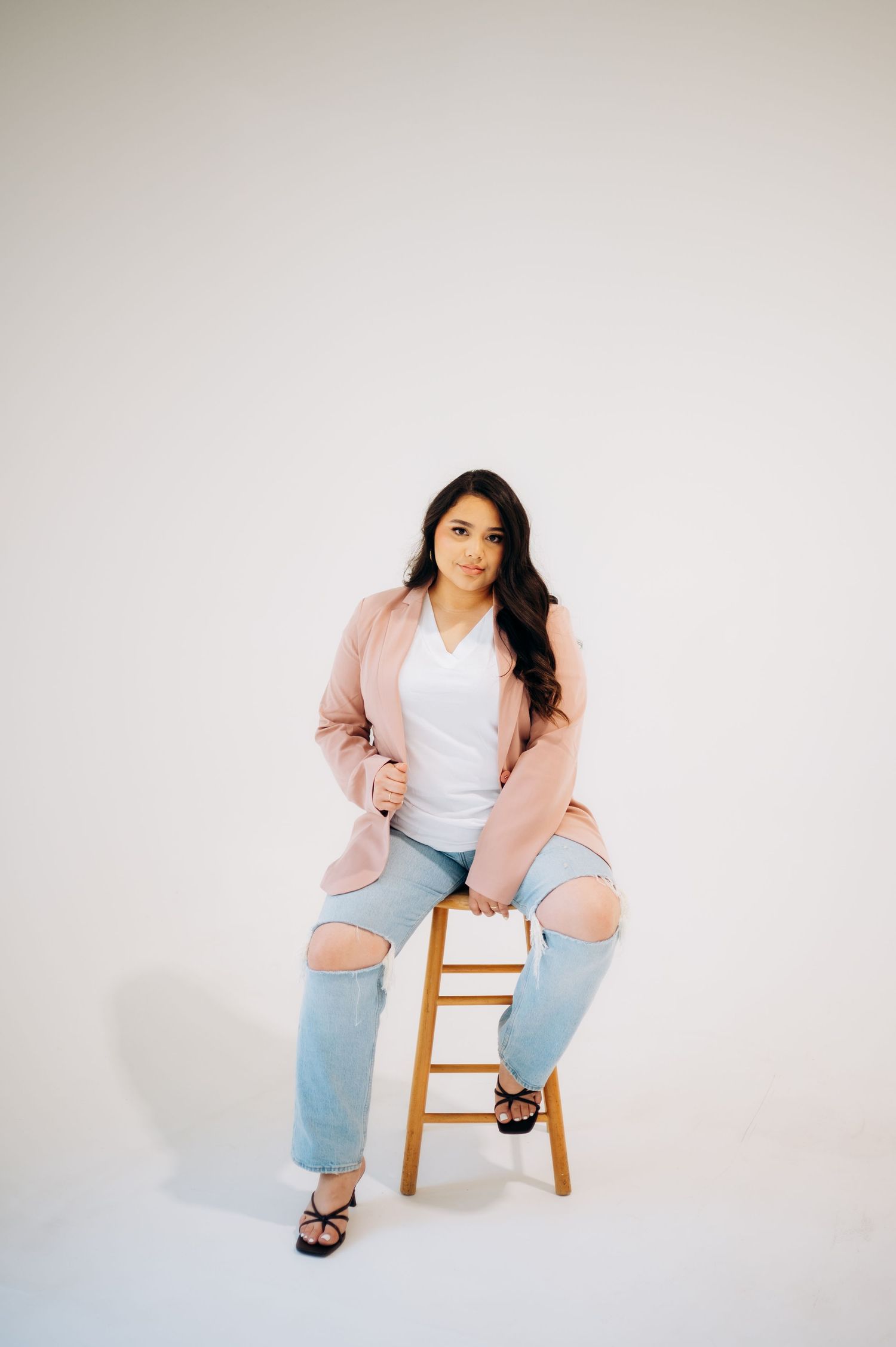 Modern fashion portrait on wooden stool featuring pink blazer, white top and distressed jeans.