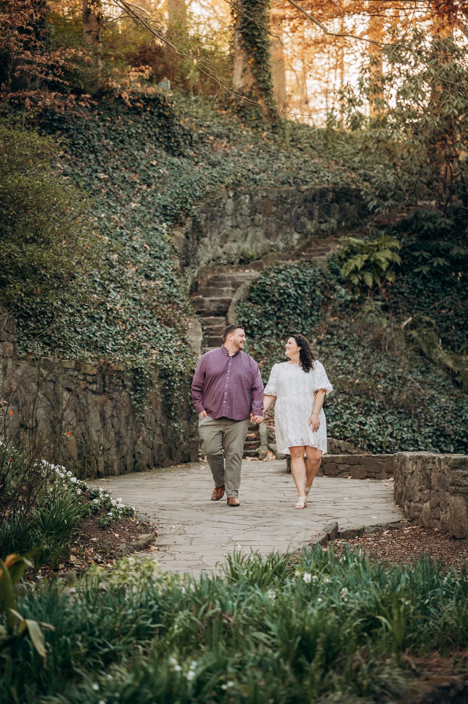 A couple walks hand in hand along a winding stone path surrounded by lush greenery and ivy-covered walls at sunset.
