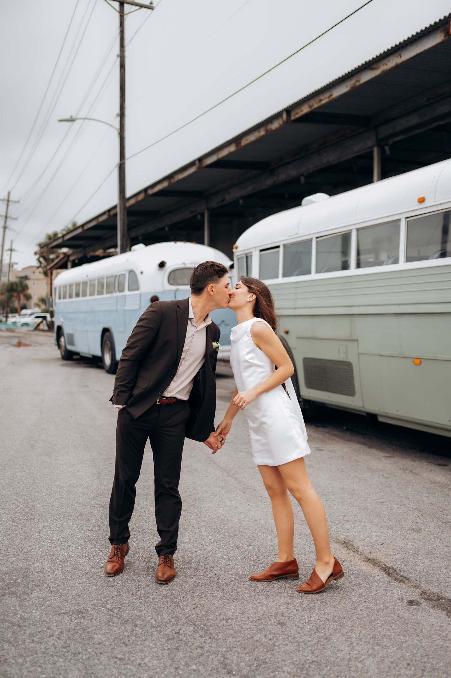 Newlyweds share a kiss while holding hands in front of vintage white buses on their wedding day.