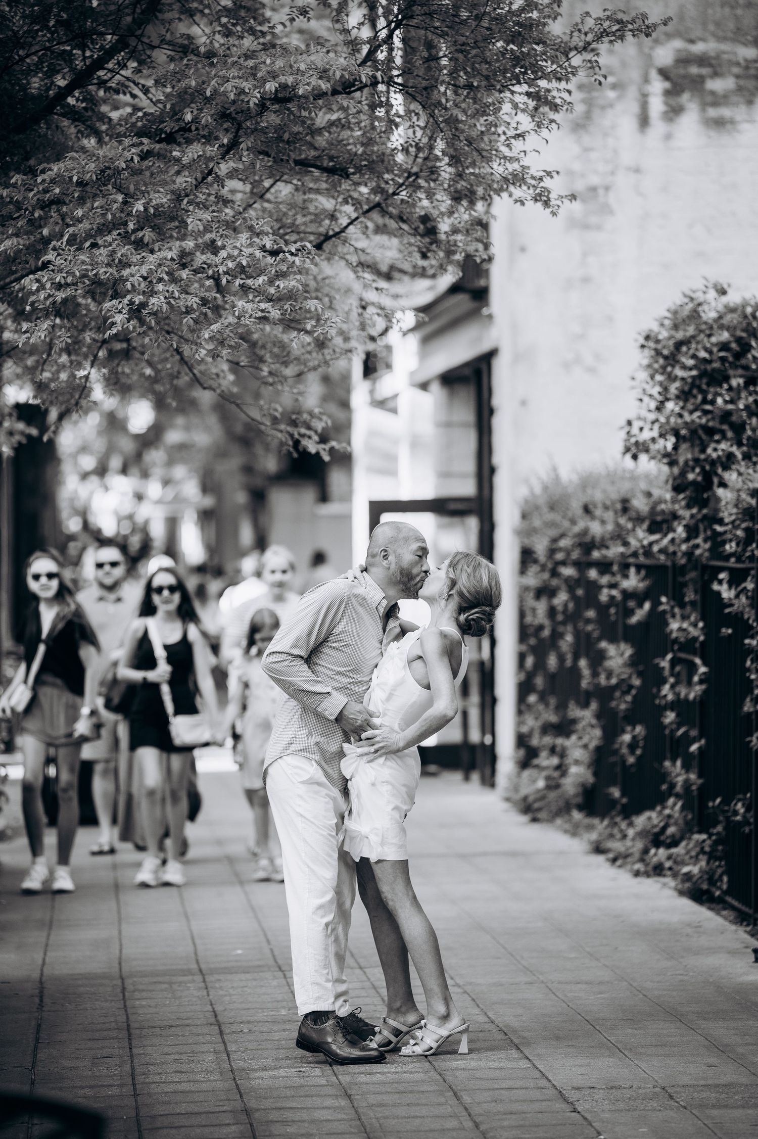 Black and white photo of couple kissing on busy city sidewalk with pedestrians walking in background.