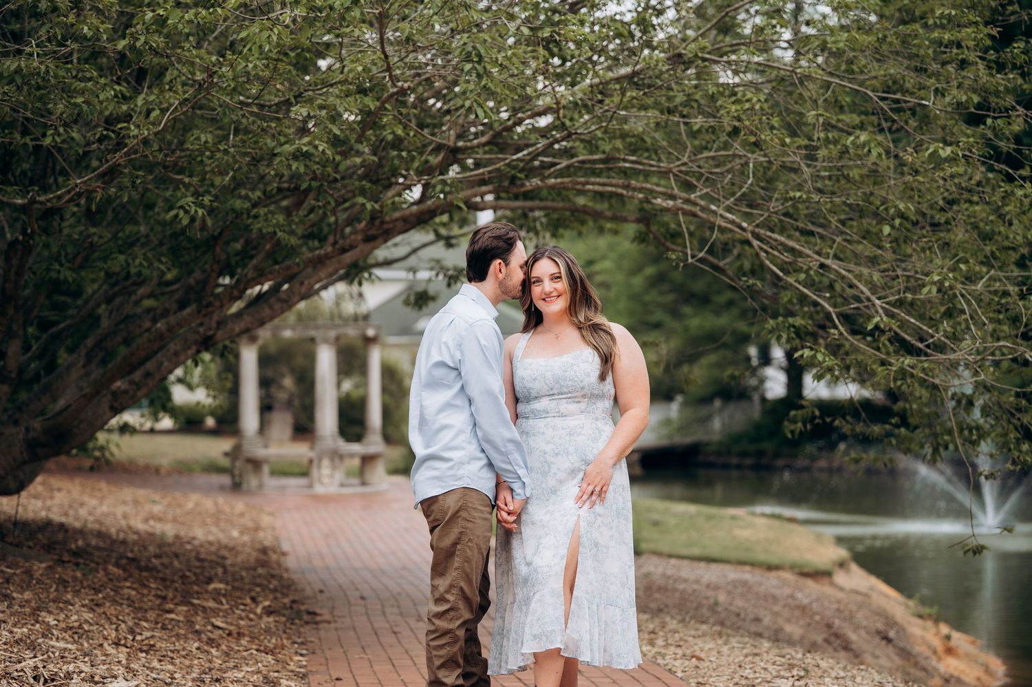 Couple holding hands and walking on brick path with man kissing woman near fountain.