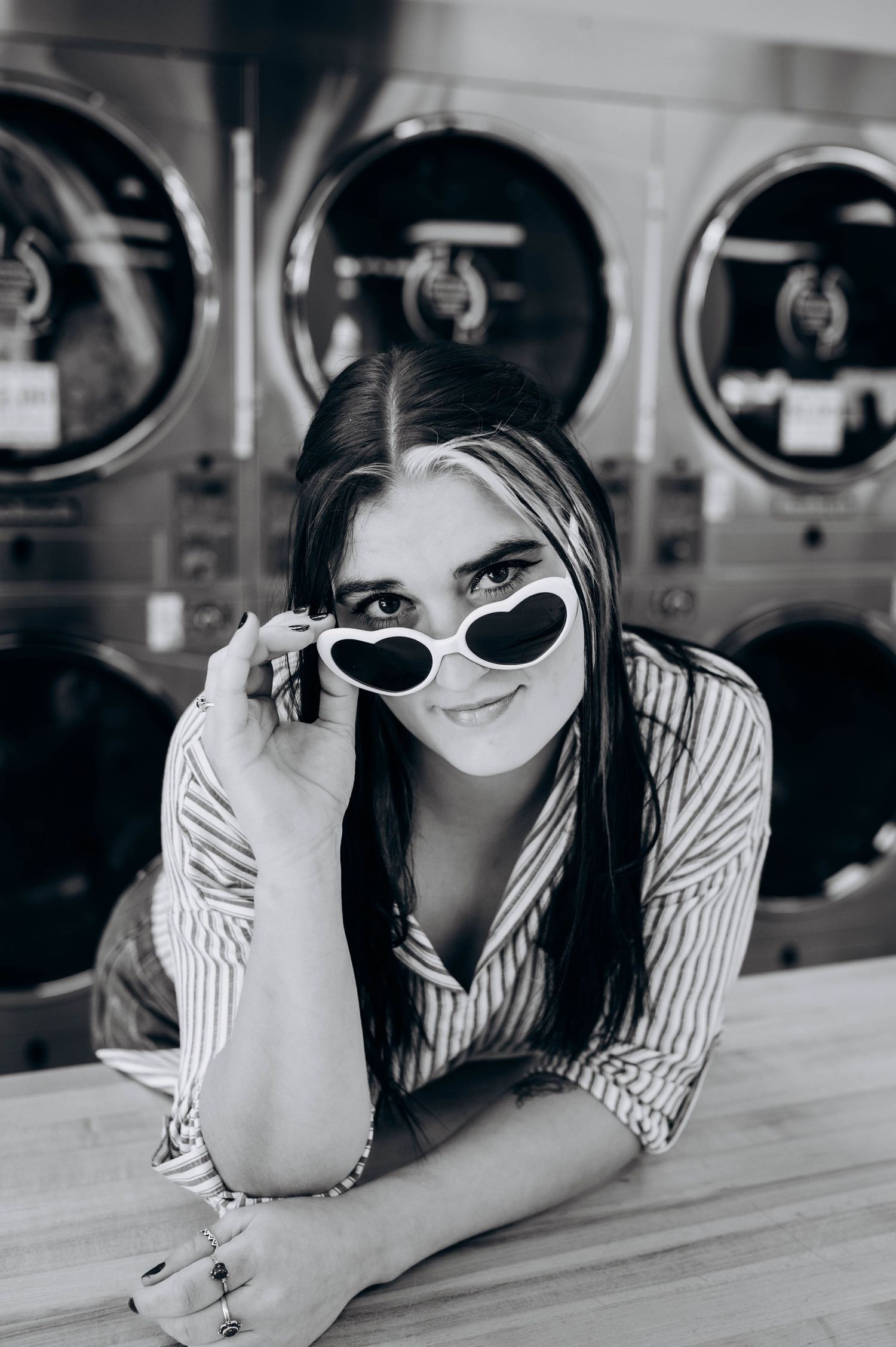 Black and white photo of someone in heart-shaped sunglasses and striped shirt at laundromat counter.