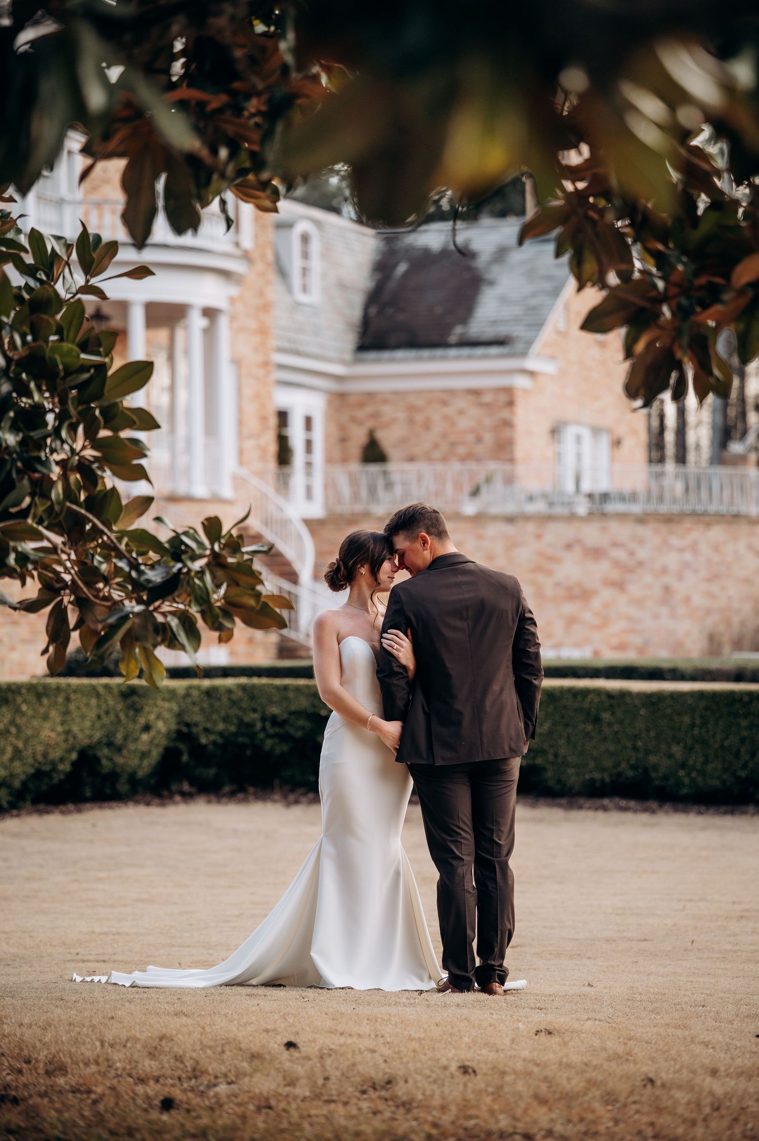 Wedding couple shares intimate moment in garden setting with magnolia trees and brick mansion backdrop.