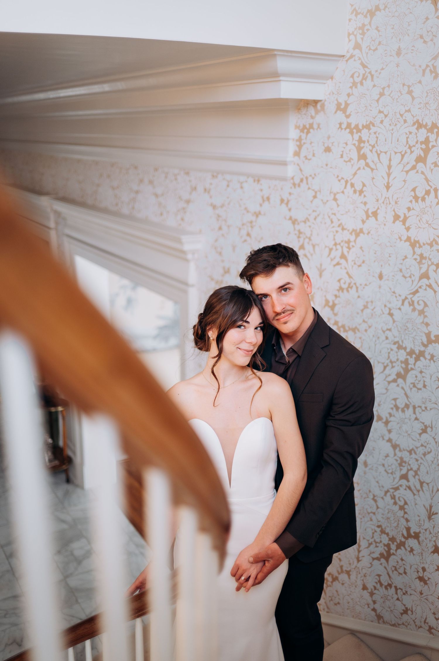 Couple poses on elegant staircase with decorative wallpaper background.
