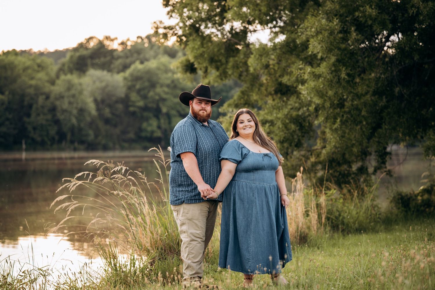 Couple holding hands near a serene lake at golden hour, man in cowboy hat and woman in blue dress.