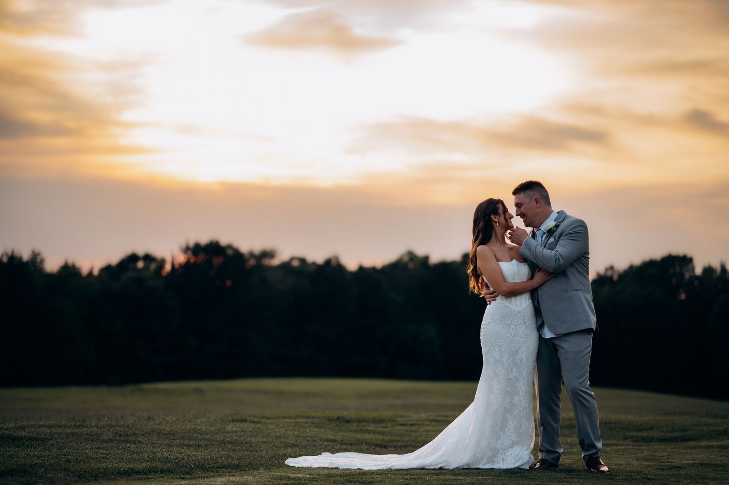 Bride and groom embrace at sunset on a lush green field, golden sky glowing behind them.