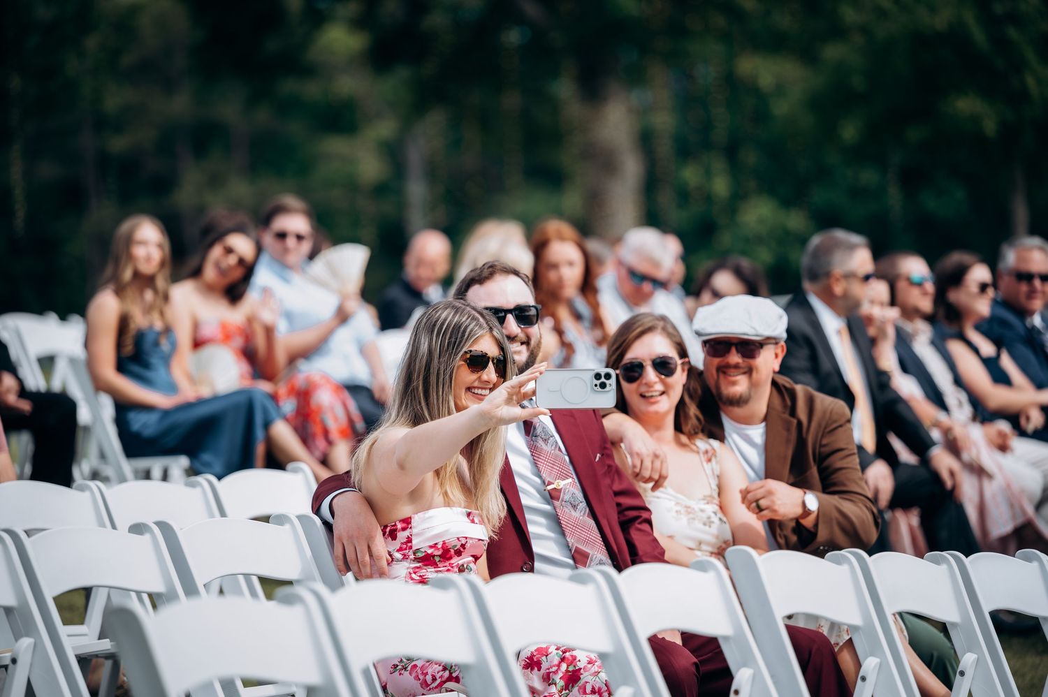 Wedding guests take selfie while seated in white chairs at outdoor ceremony in wooded setting.