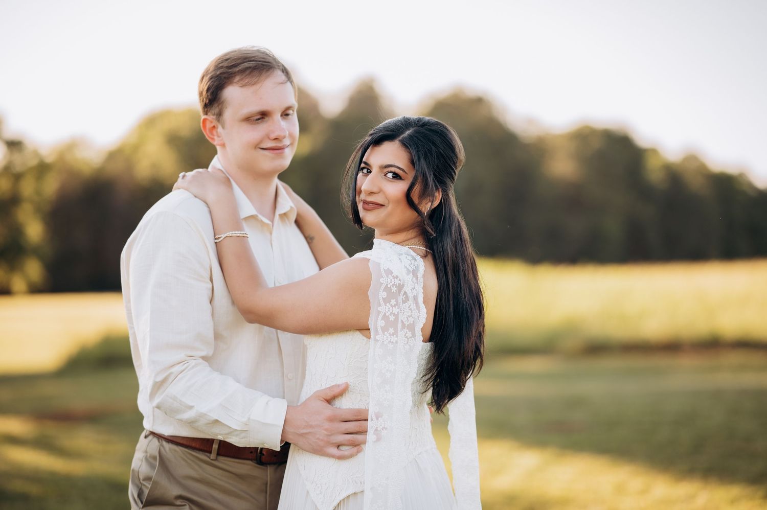 Couple embracing in golden hour light in a green field, woman in white lace dress, man in beige shirt.