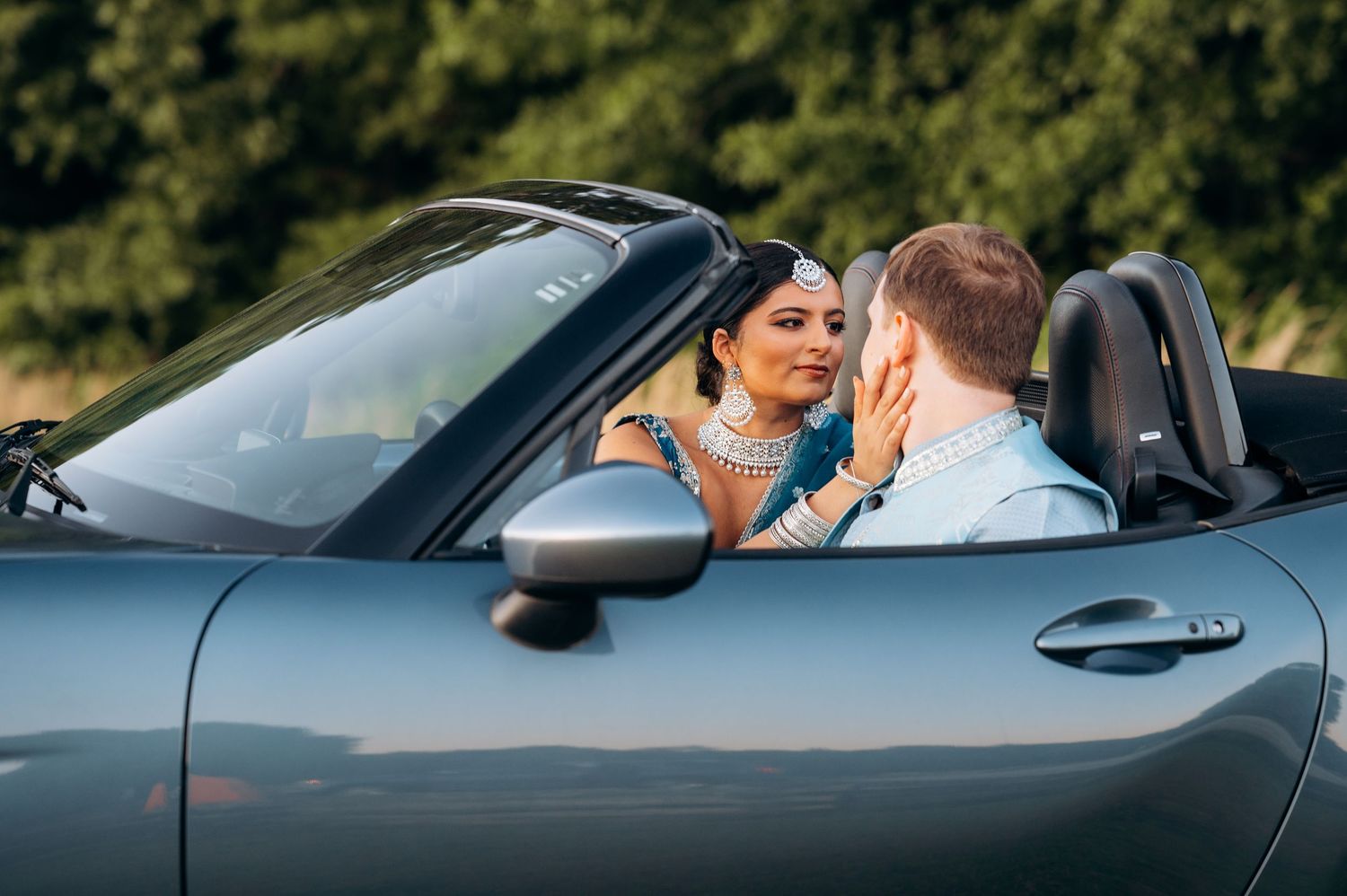 Couple in traditional Indian bridal attire sharing a romantic moment in a convertible sports car outdoors.