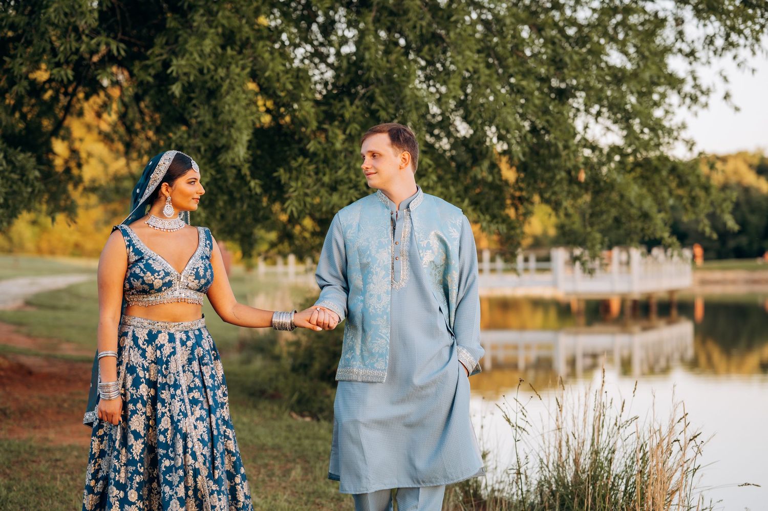 Couple in traditional Indian attire holding hands outdoors near a pond, woman in blue lehenga, man in blue kurta.