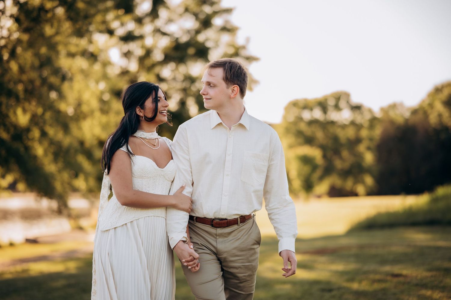 Couple in white and beige attire holding hands and walking through a lush green park during golden hour.