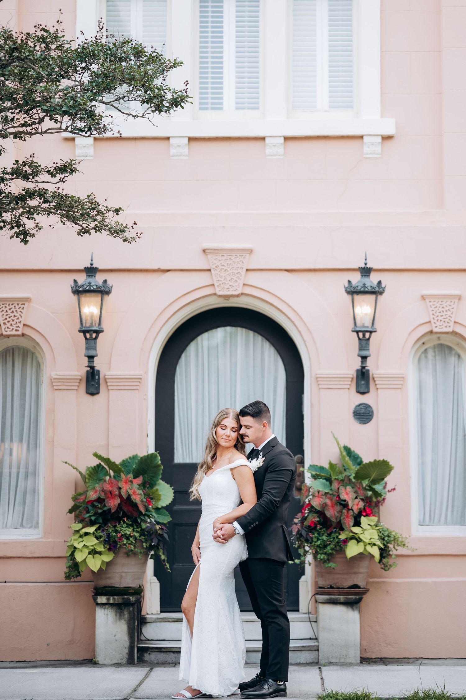 Bride and groom embracing in front of a pink building with arched door and lush floral arrangements in Charleston, SC.