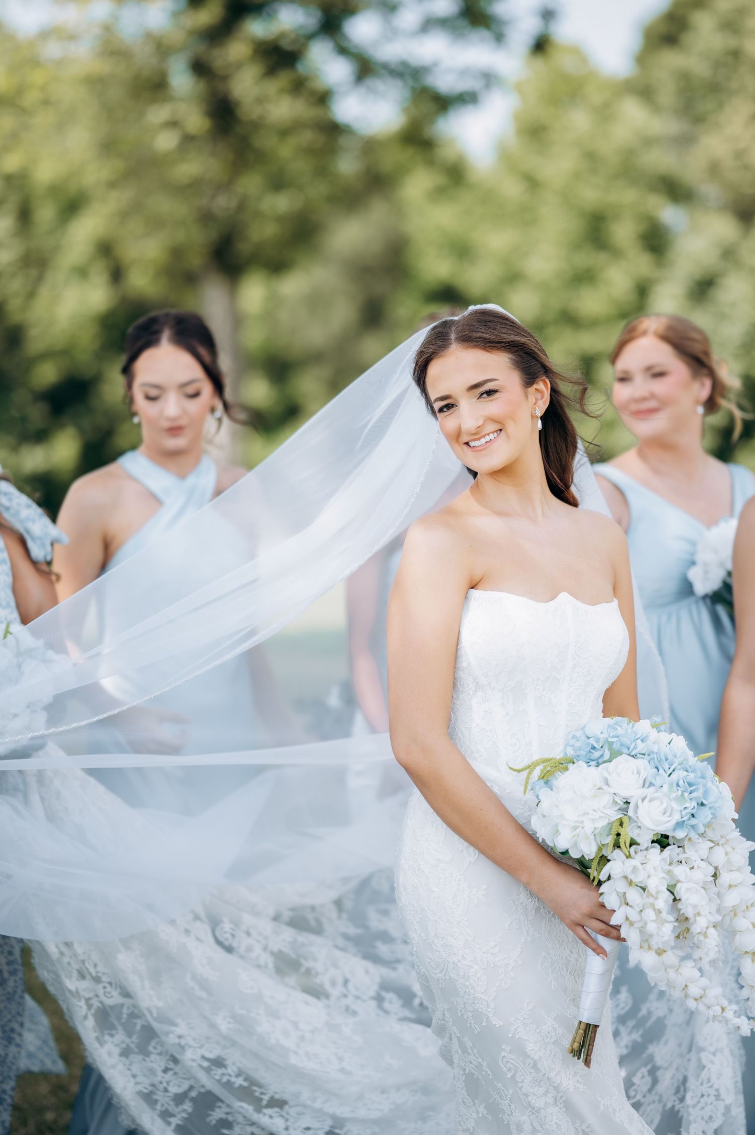 Smiling bride in strapless lace gown holds blue floral bouquet as bridesmaids in light blue dresses hold her veil outdoors.