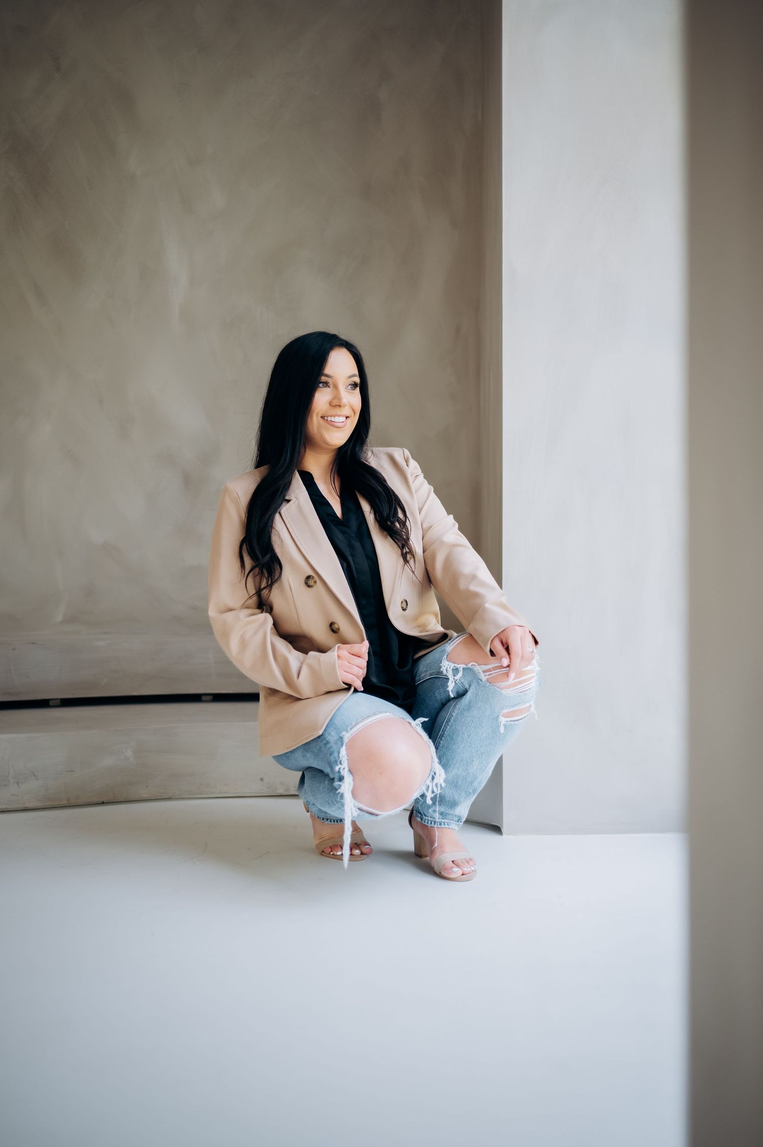 Stylish woman in beige blazer and ripped jeans crouches against neutral wall in minimalist indoor setting.