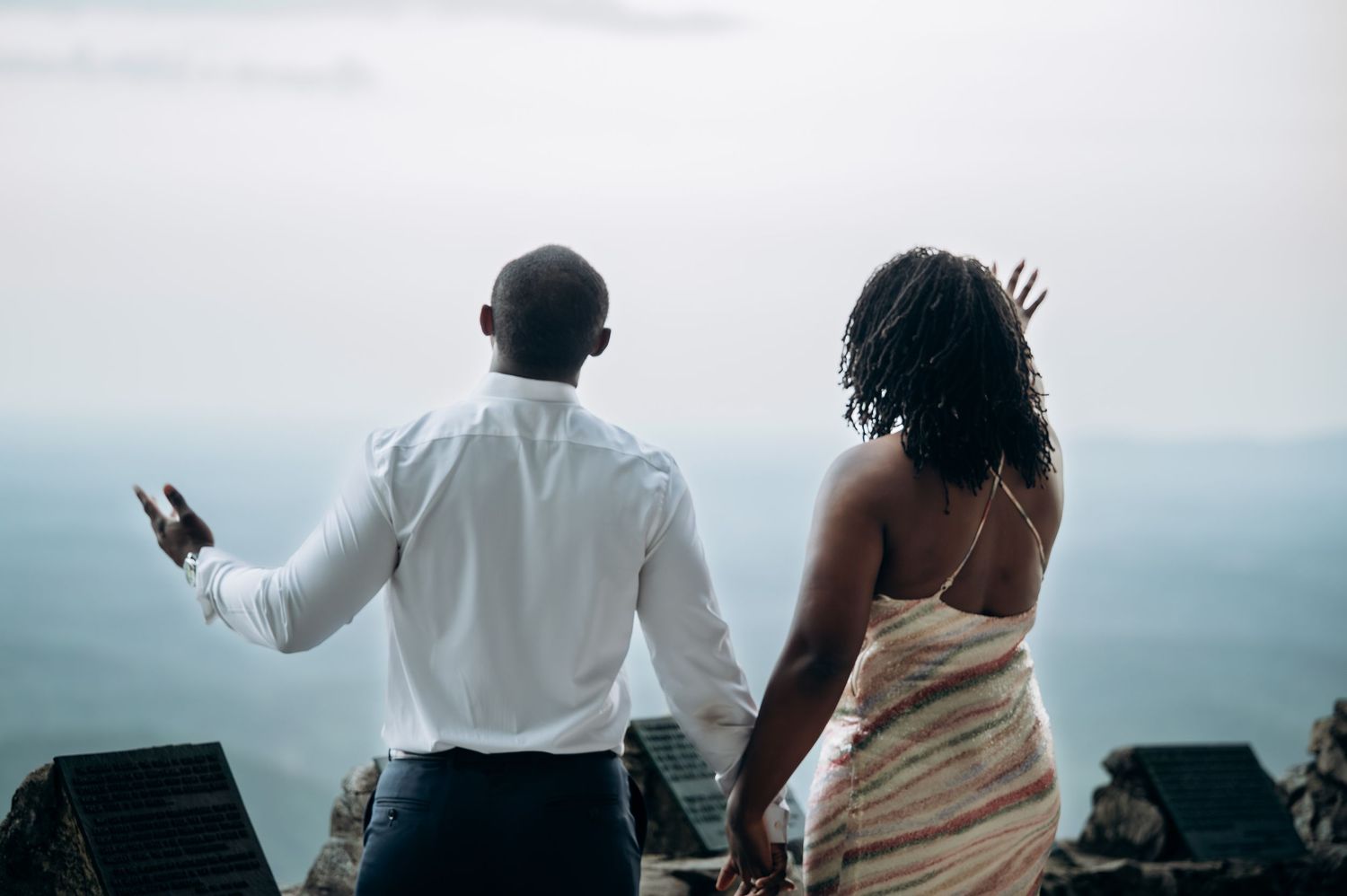 A couple stands back-to-back on a scenic overlook, the man in white shirt gesturing toward a misty landscape.
