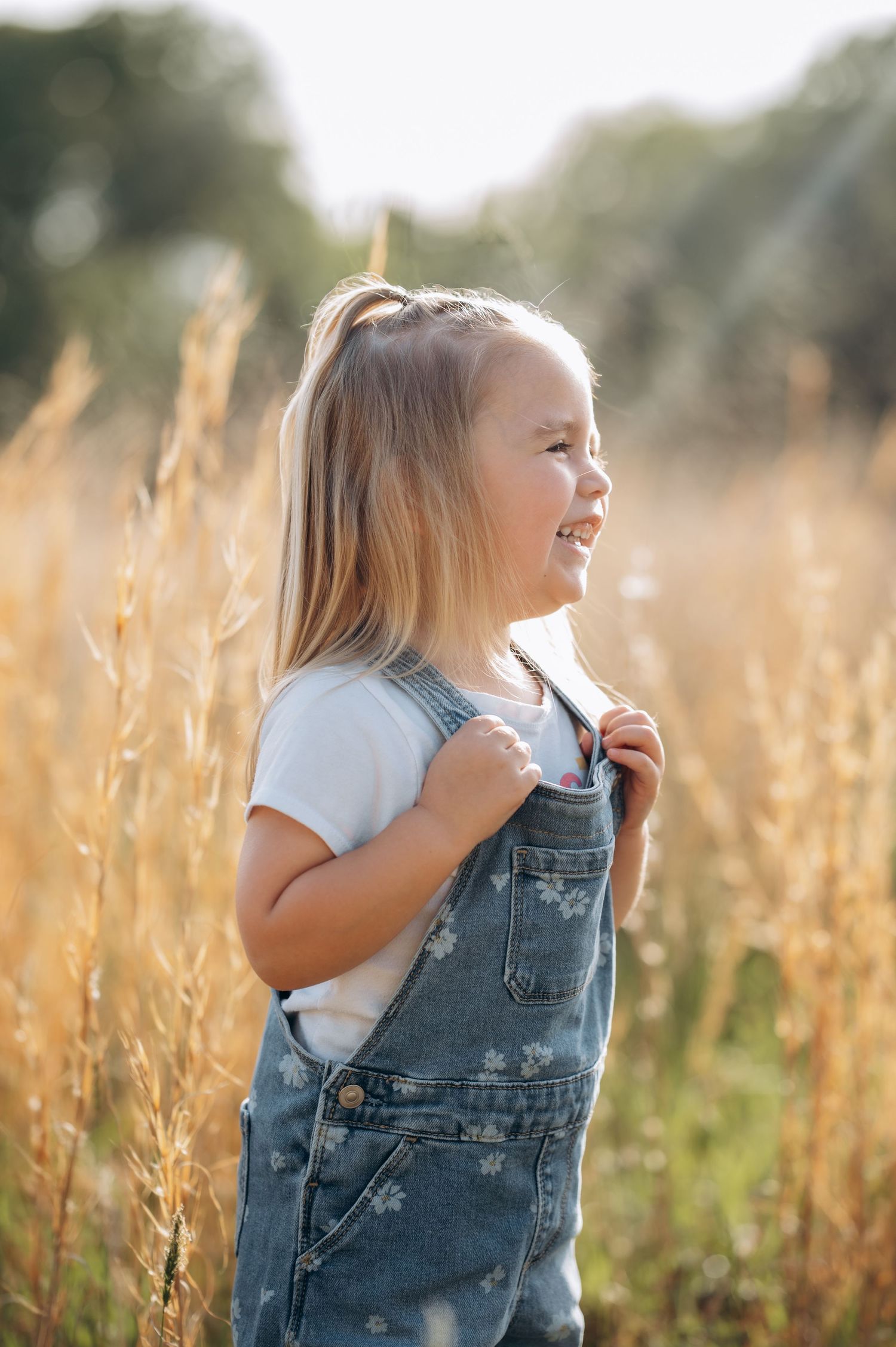A smiling little girl in denim overalls stands in a golden field, enjoying a sunny day outdoors.