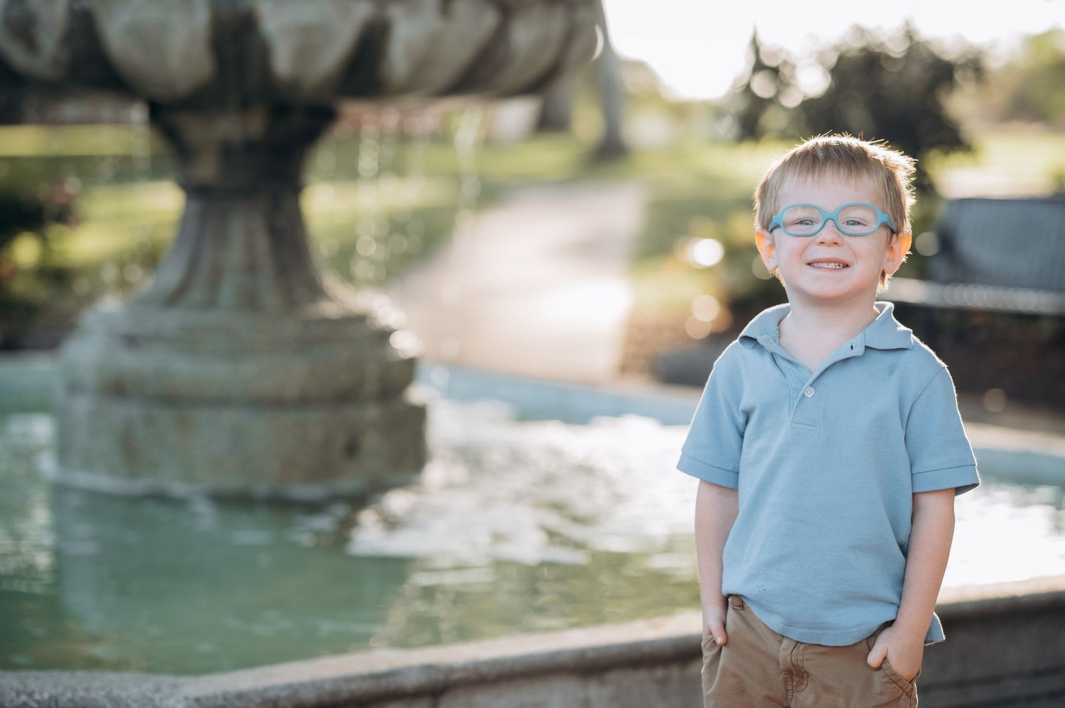 Smiling young boy with glasses in a light blue polo shirt standing near a decorative stone fountain outdoors.
