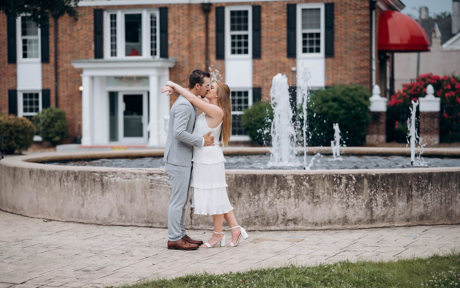 Couple in white and grey attire posing near fountain outside traditional brick building.