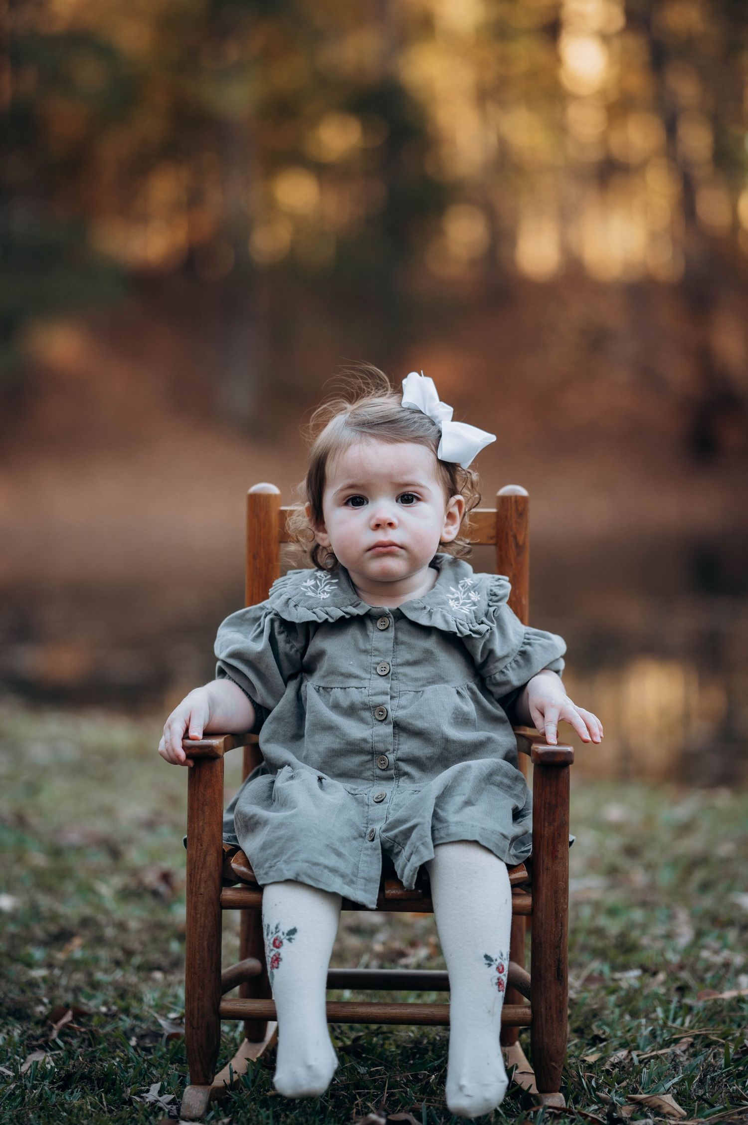 A toddler in a gray dress and white bow sits in a wooden rocking chair outdoors.