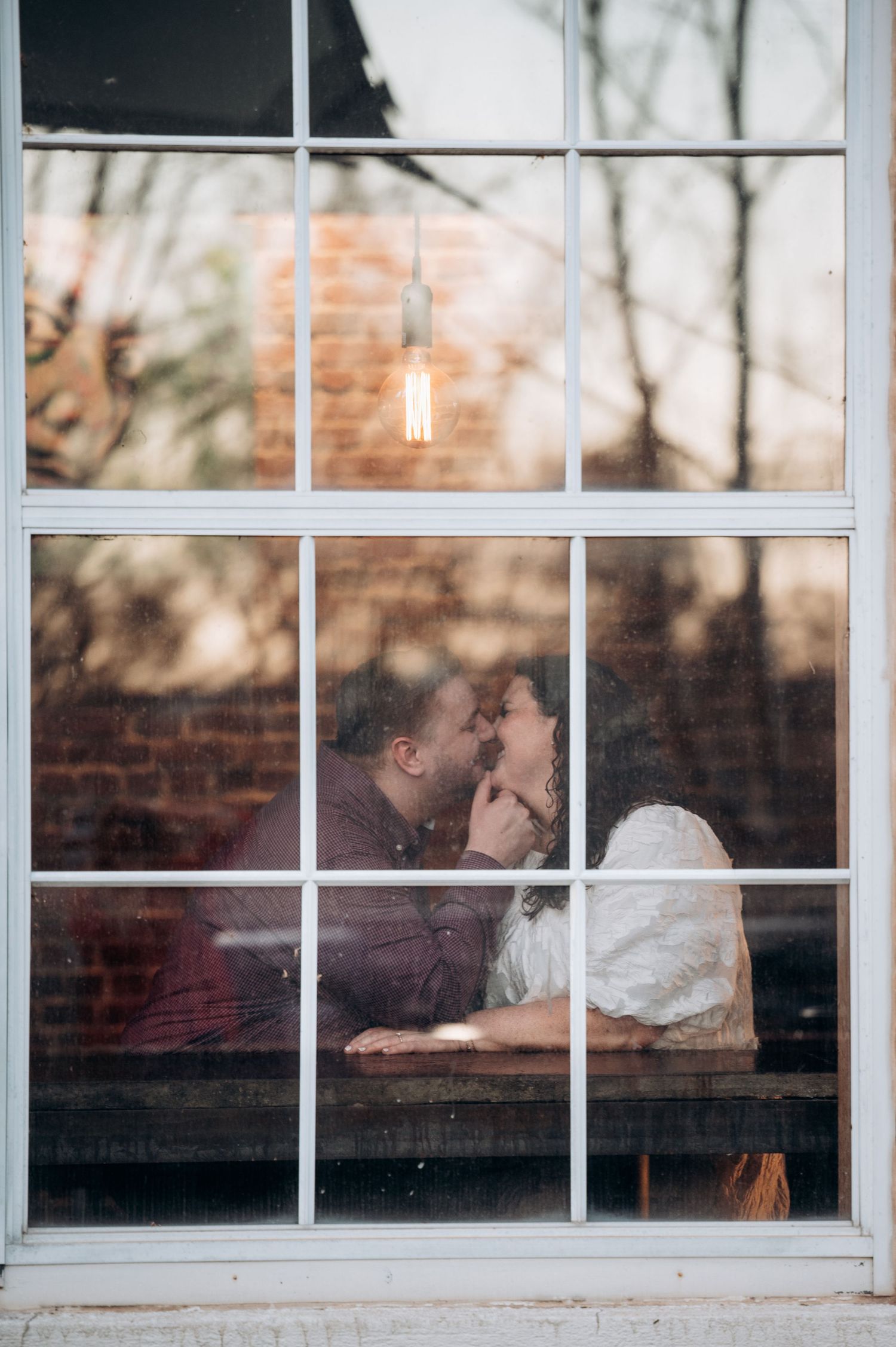 A romantic couple embraces near a window with white panes, creating a dreamy silhouette against natural light.