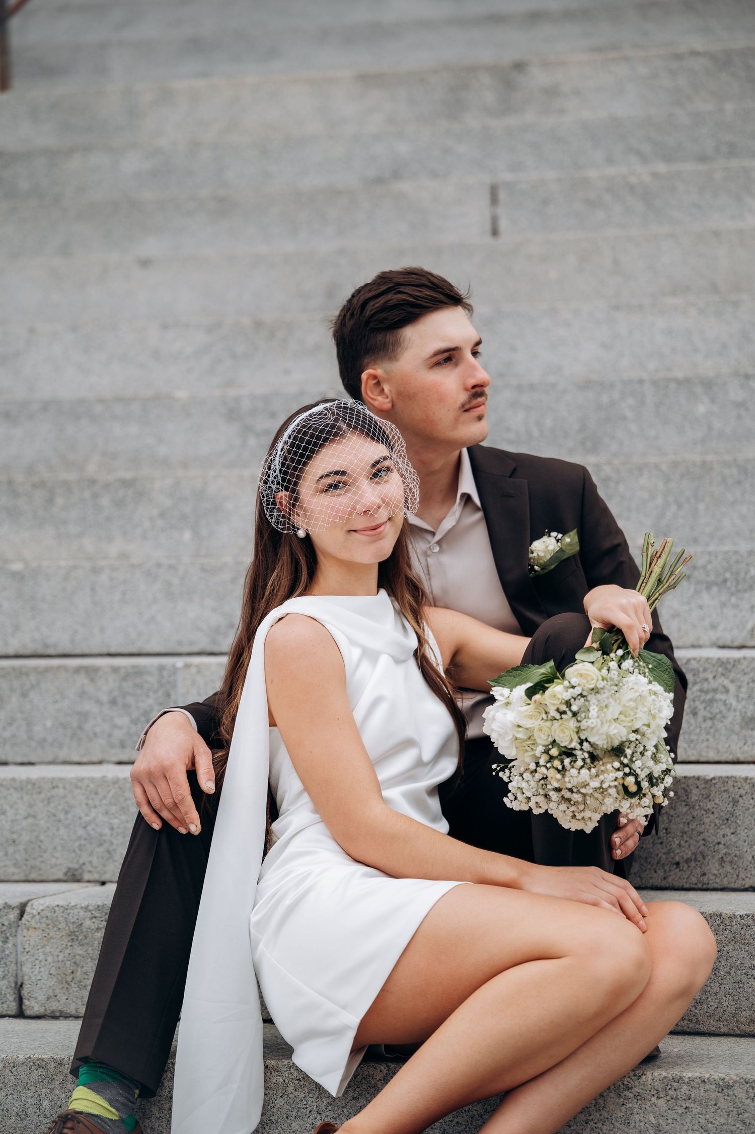 Couple sits intimately on concrete steps with white flowers.
