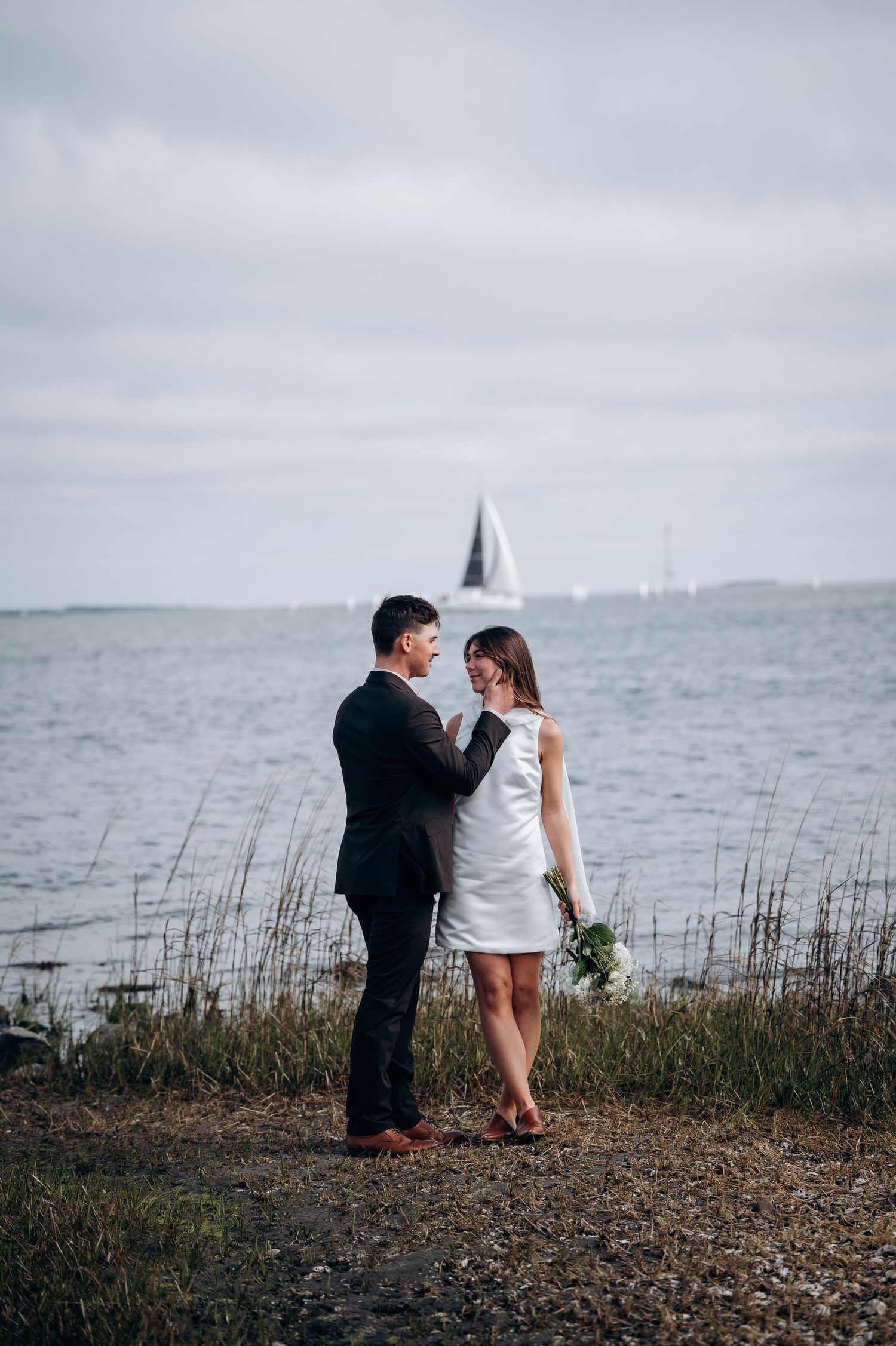 A couple shares an intimate moment against a coastal backdrop with tall grass and water views.