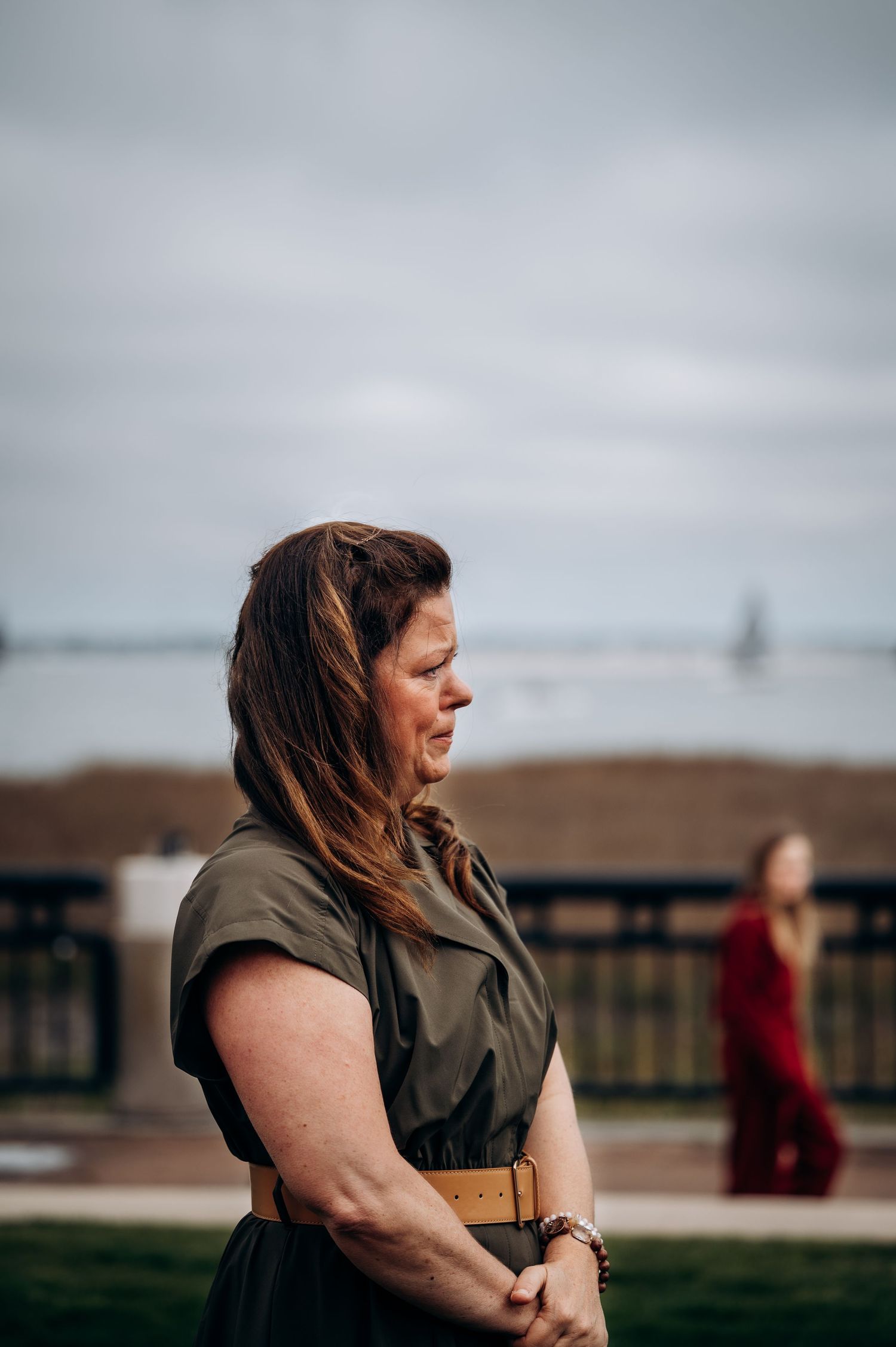 Woman in olive dress standing outdoors near waterfront with wind blowing her brown hair.