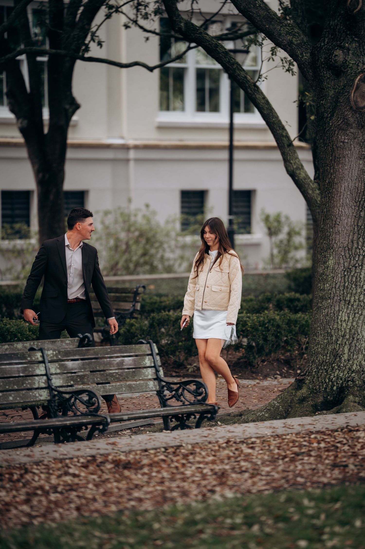 Couple walking together through a tree-lined park path with benches on an overcast day.