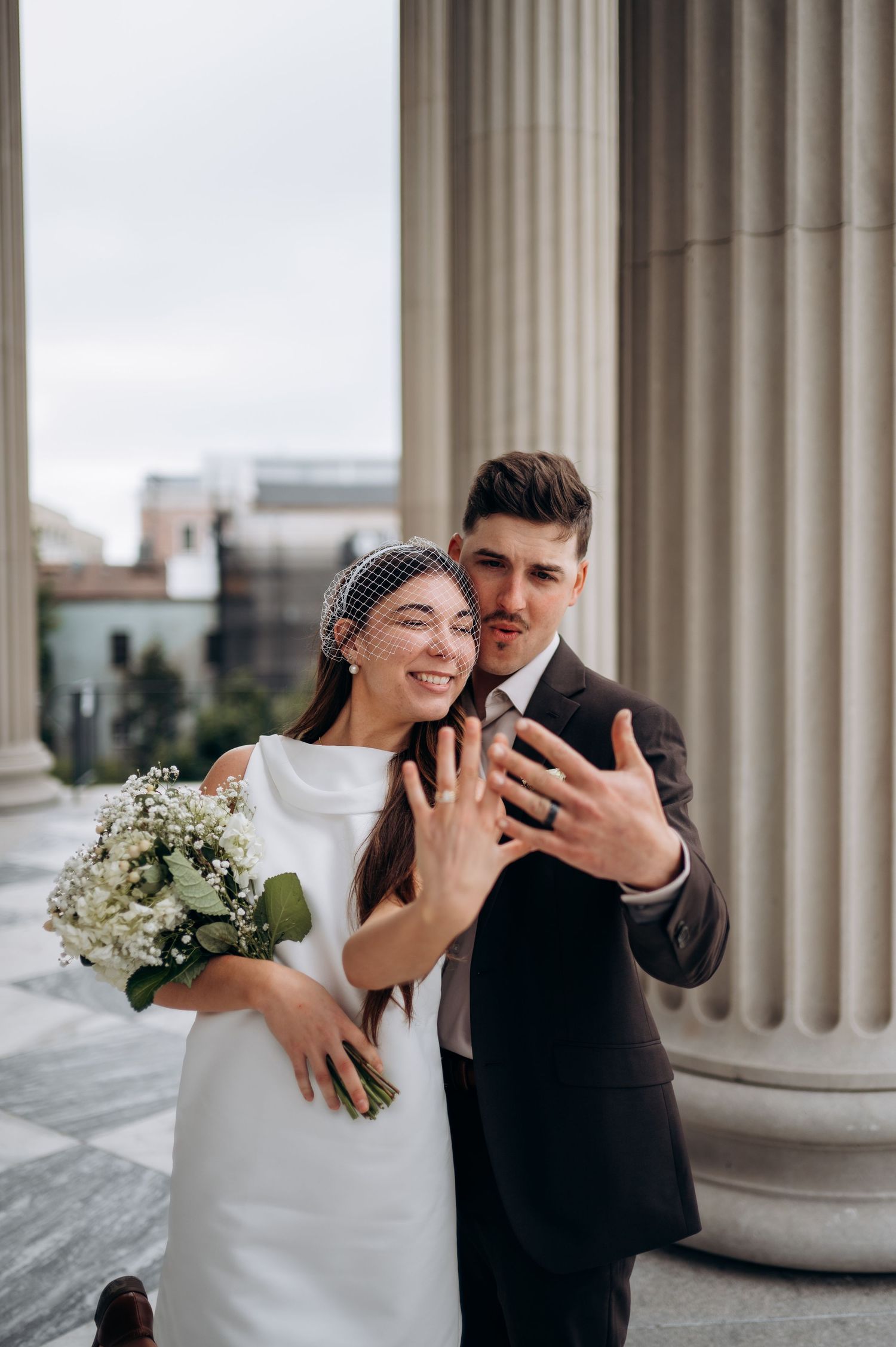 Newlyweds showing off wedding rings while smiling, bride in white dress with birdcage veil beside groom in dark suit.