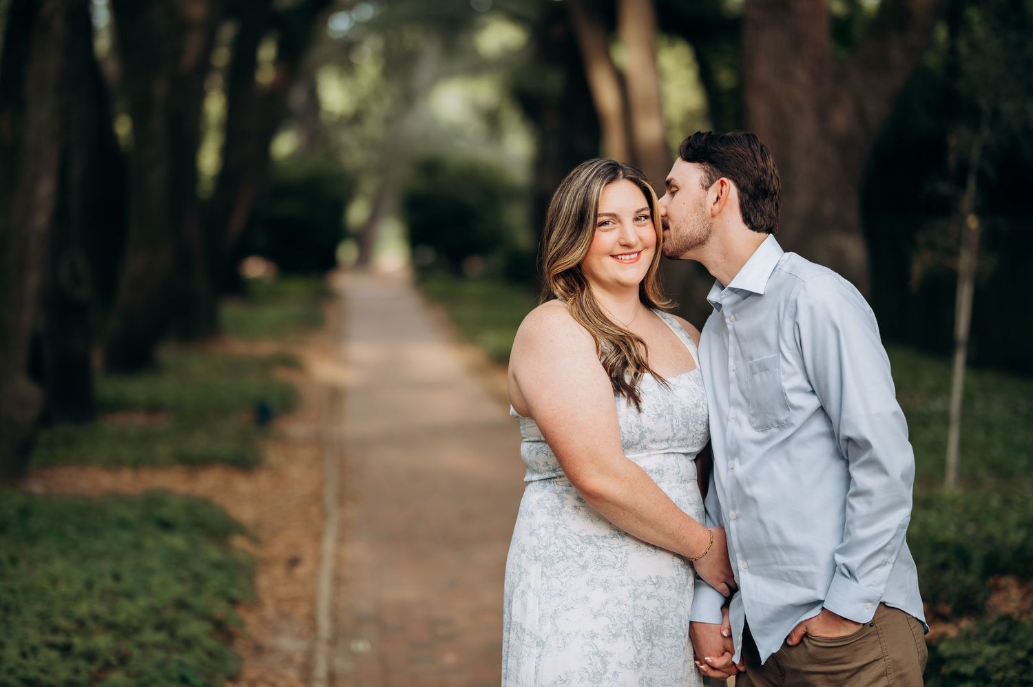 Couple sharing a romantic moment on a tree-lined path, woman in white dress smiling at camera.