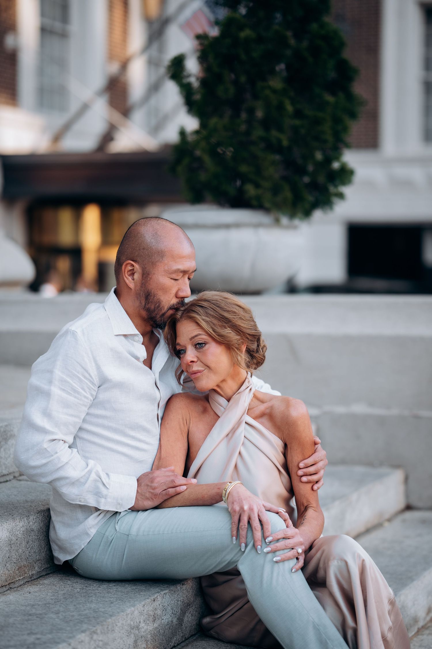 Affectionate couple sitting on stone steps, man kissing womans head while embracing outdoors.