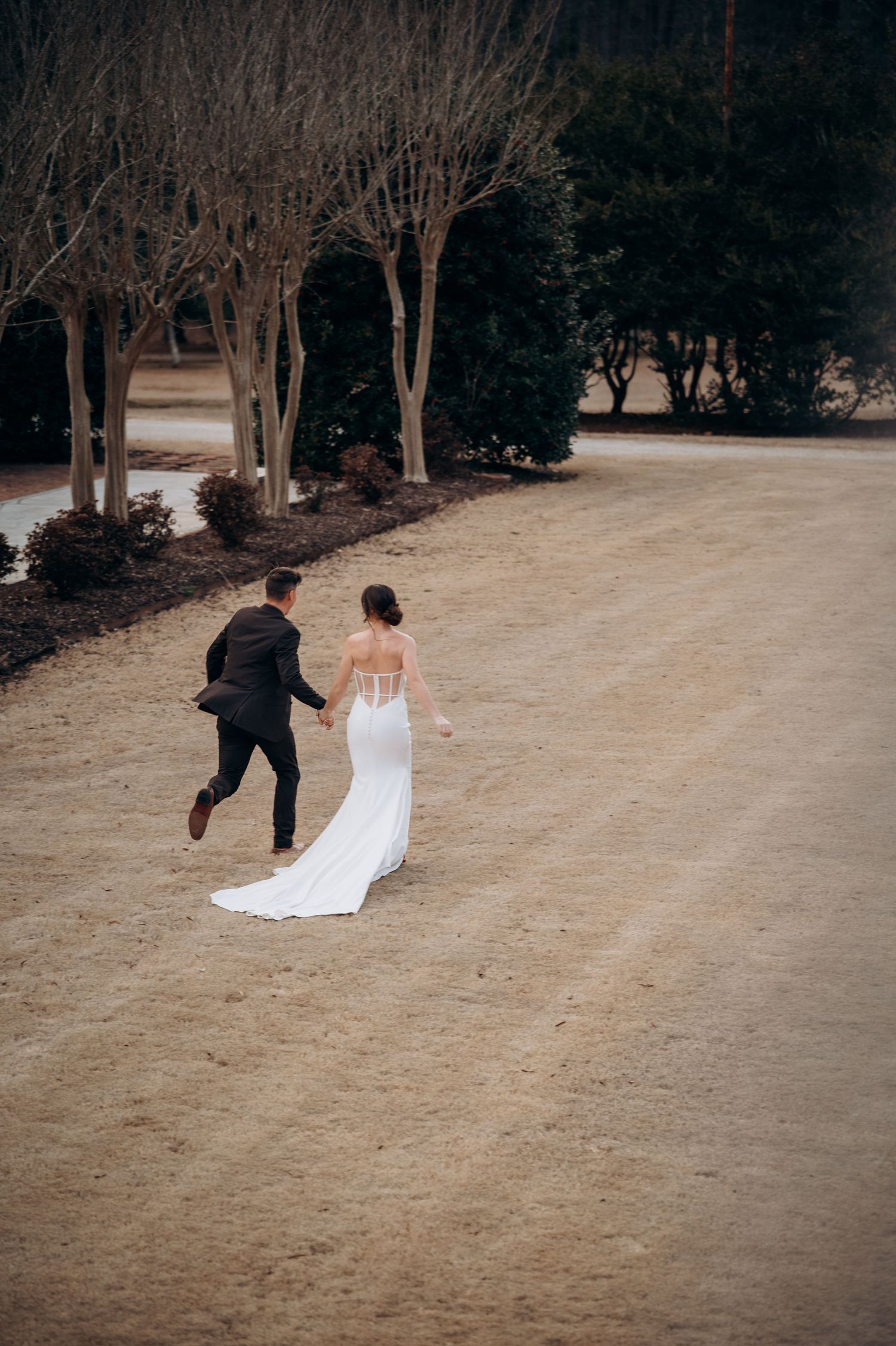 Bride in elegant white gown and groom in black suit walk hand in hand across a sandy outdoor venue.
