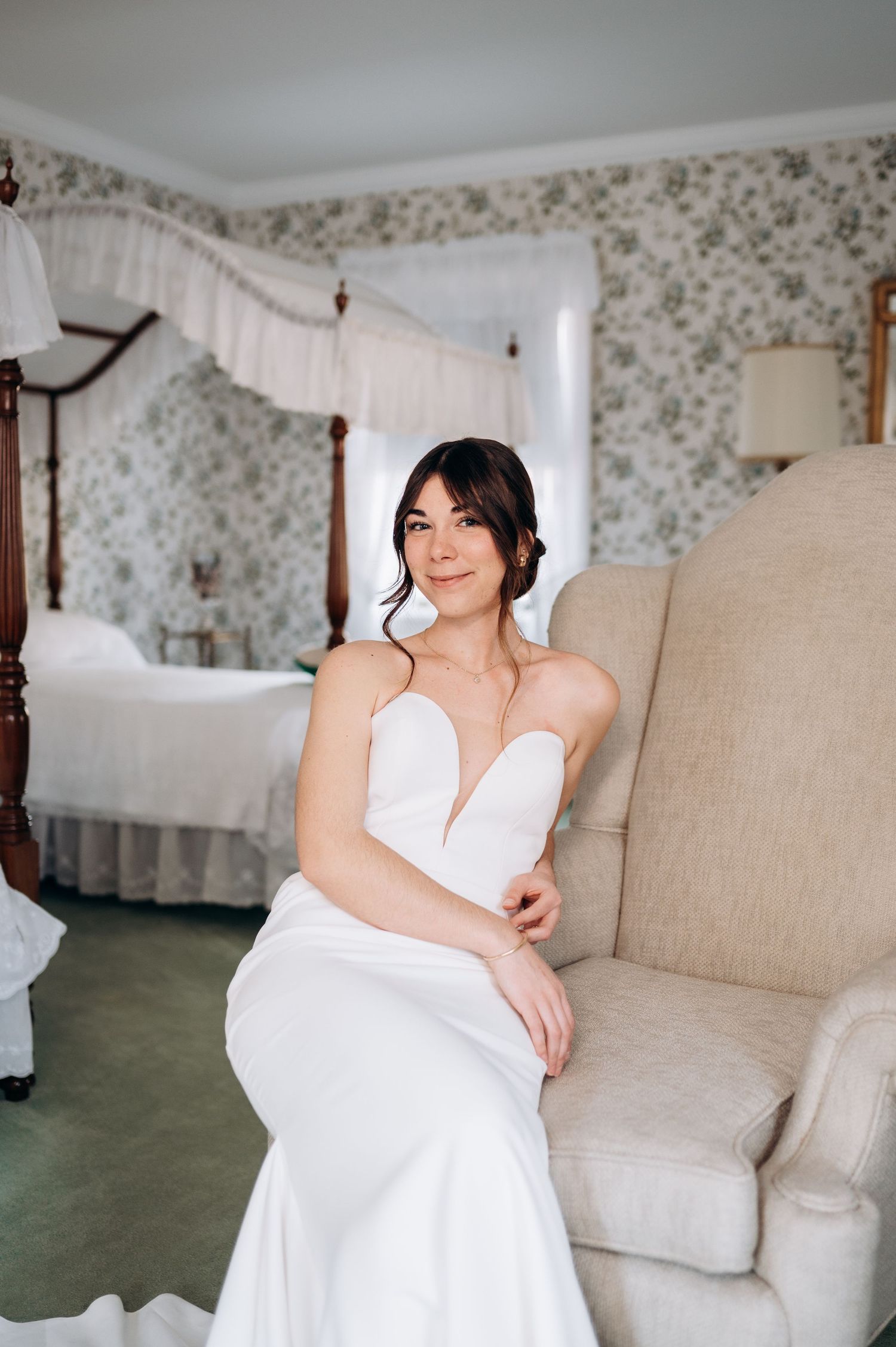 Elegant bride in strapless white mermaid gown seated in armchair in floral wallpapered bridal suite.