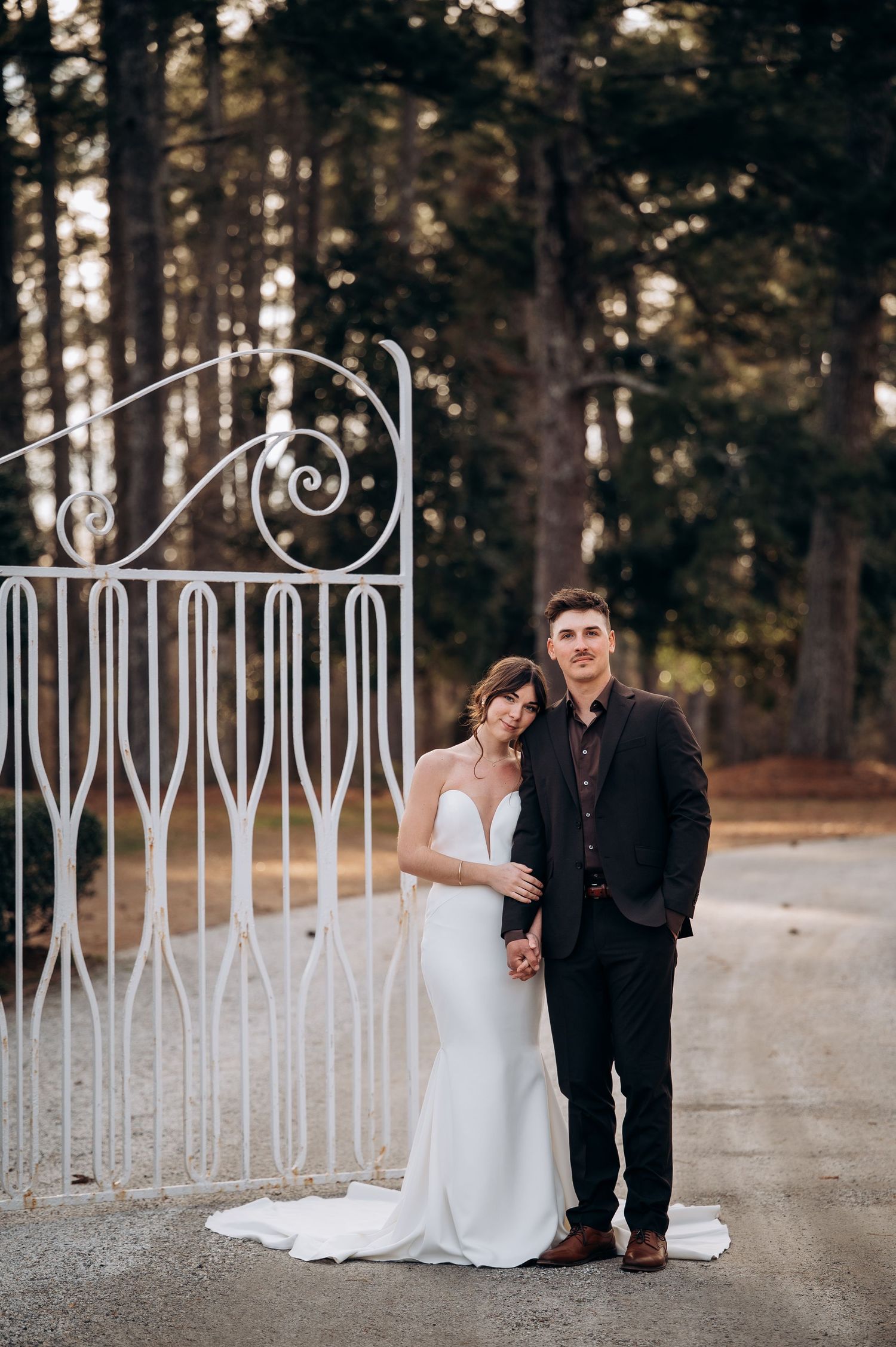 Bride and groom posing elegantly by a white ornate iron gate surrounded by tall trees.