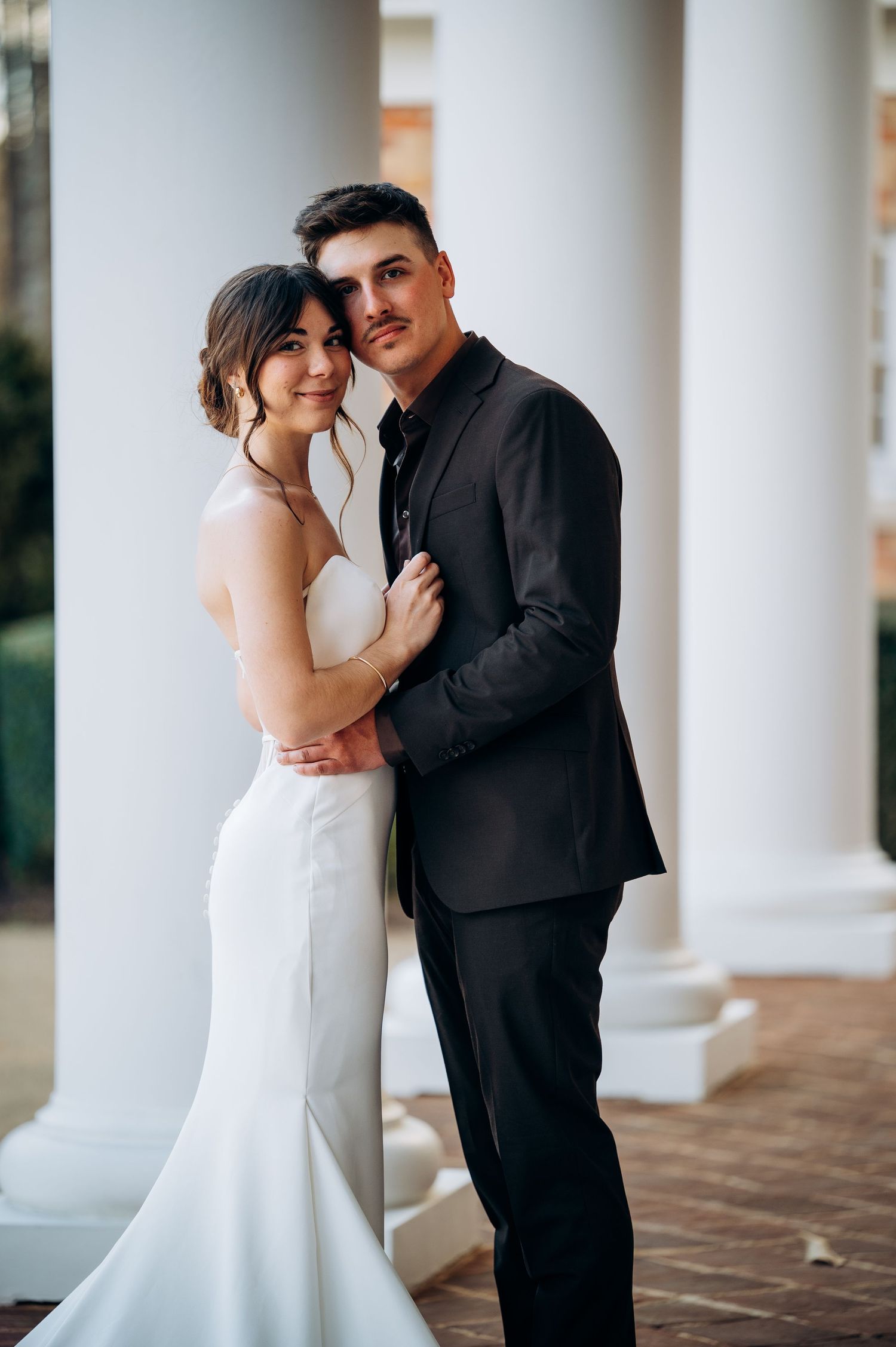 Wedding couple embracing between white columns, bride in sleek white gown, groom in black suit.