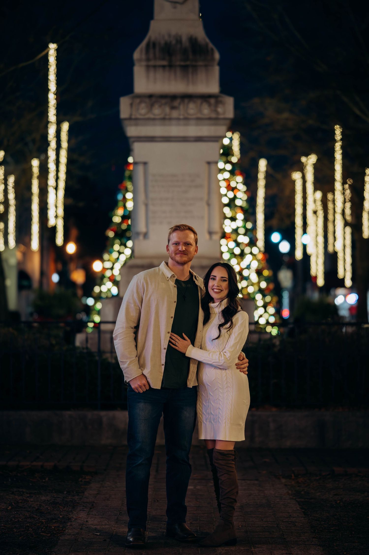 Couple posing together at night with Christmas tree lights and monument backdrop.