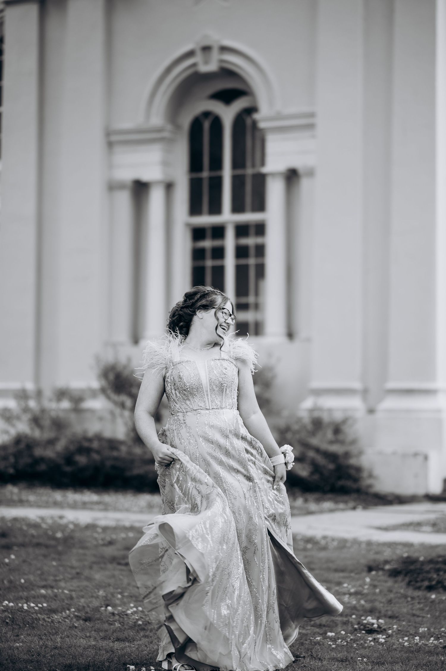 Young woman in elegant lace gown with feather details walks outdoors near white building in black and white photo.