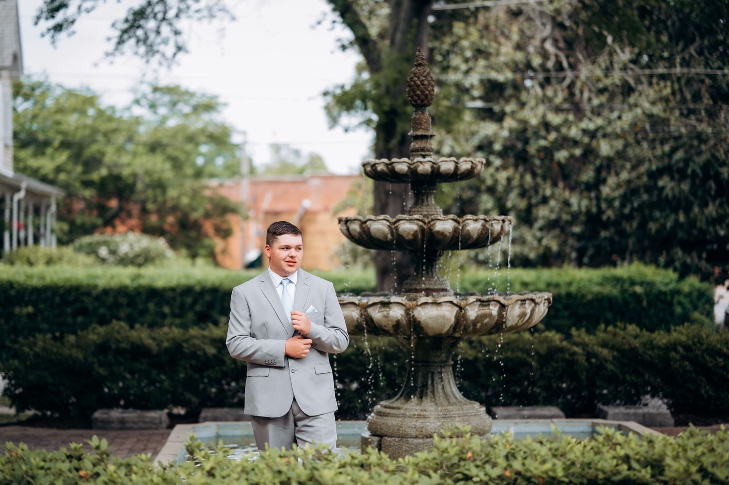 Groom in gray suit standing before elegant tiered stone fountain surrounded by lush greenery at outdoor wedding venue.